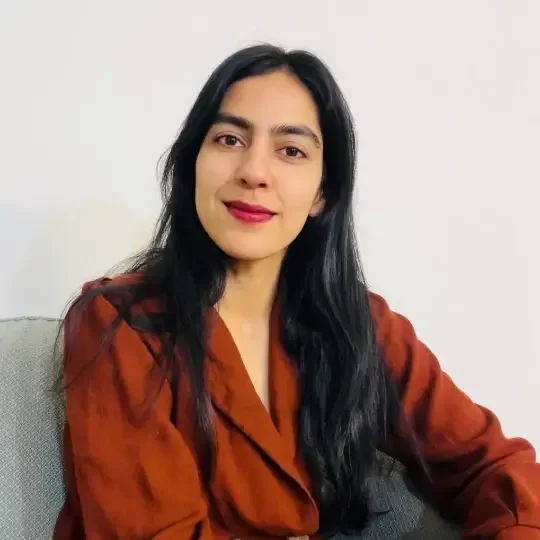 A woman with long black hair wearing a rust-colored shirt, seated on a light gray sofa against a plain white wall. | The Witch's Therapist | holistic psychotherapy in London Ontario