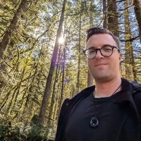 A man with glasses and a black shirt standing in a forest with tall trees and sunlight filtering through the leaves. | The Witch's Therapist | holistic psychotherapy in London Ontario