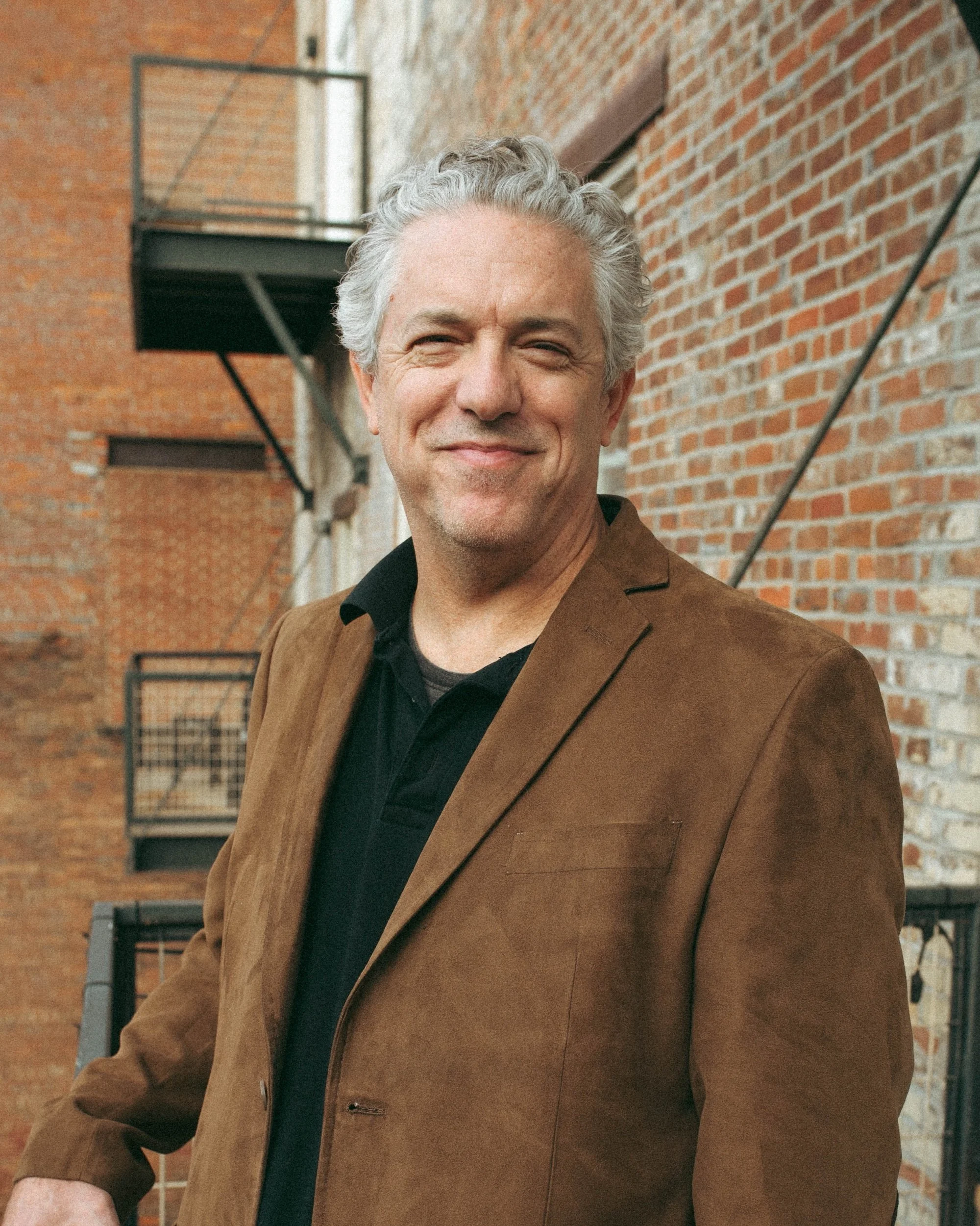 A gray-haired Gilliam Writers Group faculty member smiles before a brick wall and fire escape.