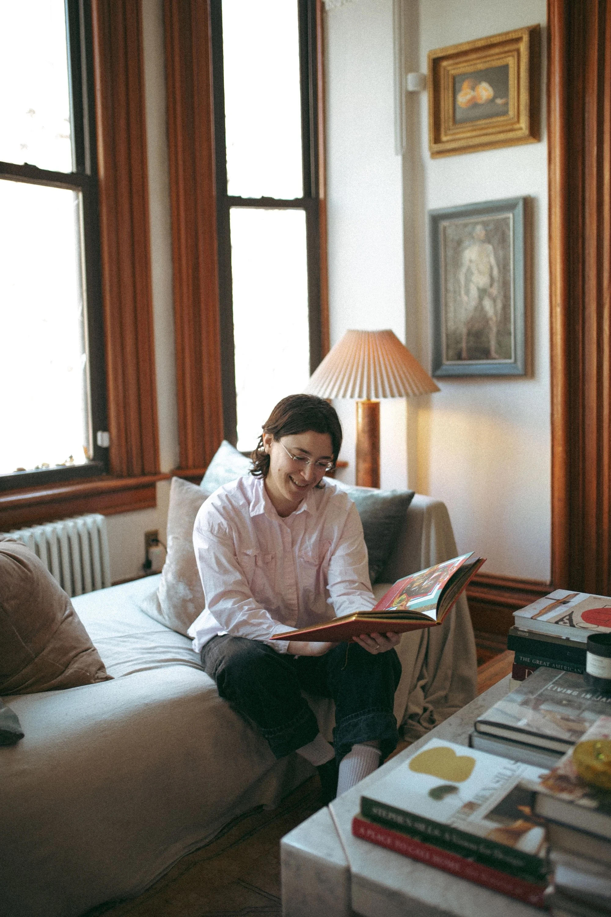 A GWG writing coach smiling over a book in a sunlit prewar living room.