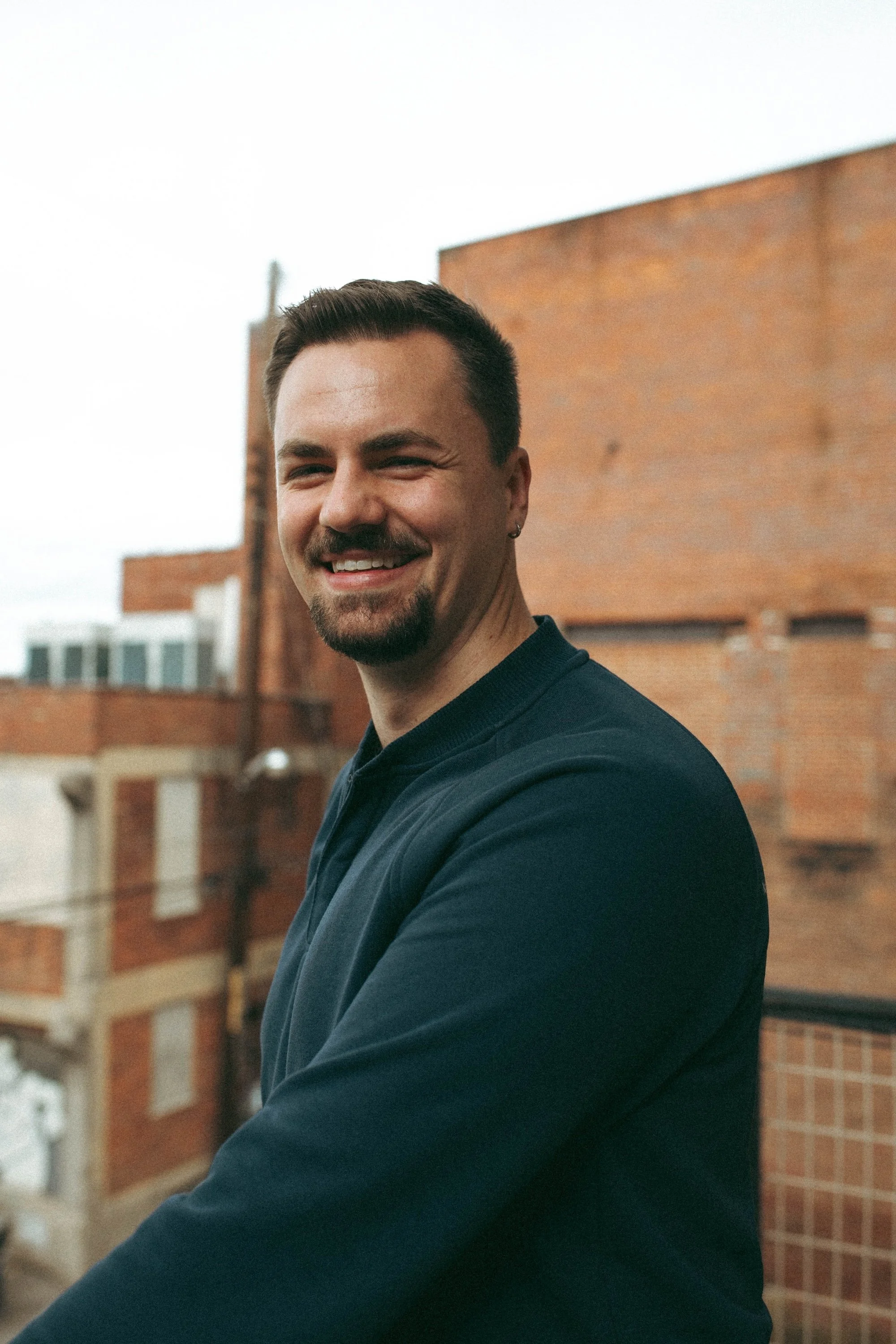 A smiling Gilliam Writers Group faculty member poses outdoors against weathered brick rooftop buildings.