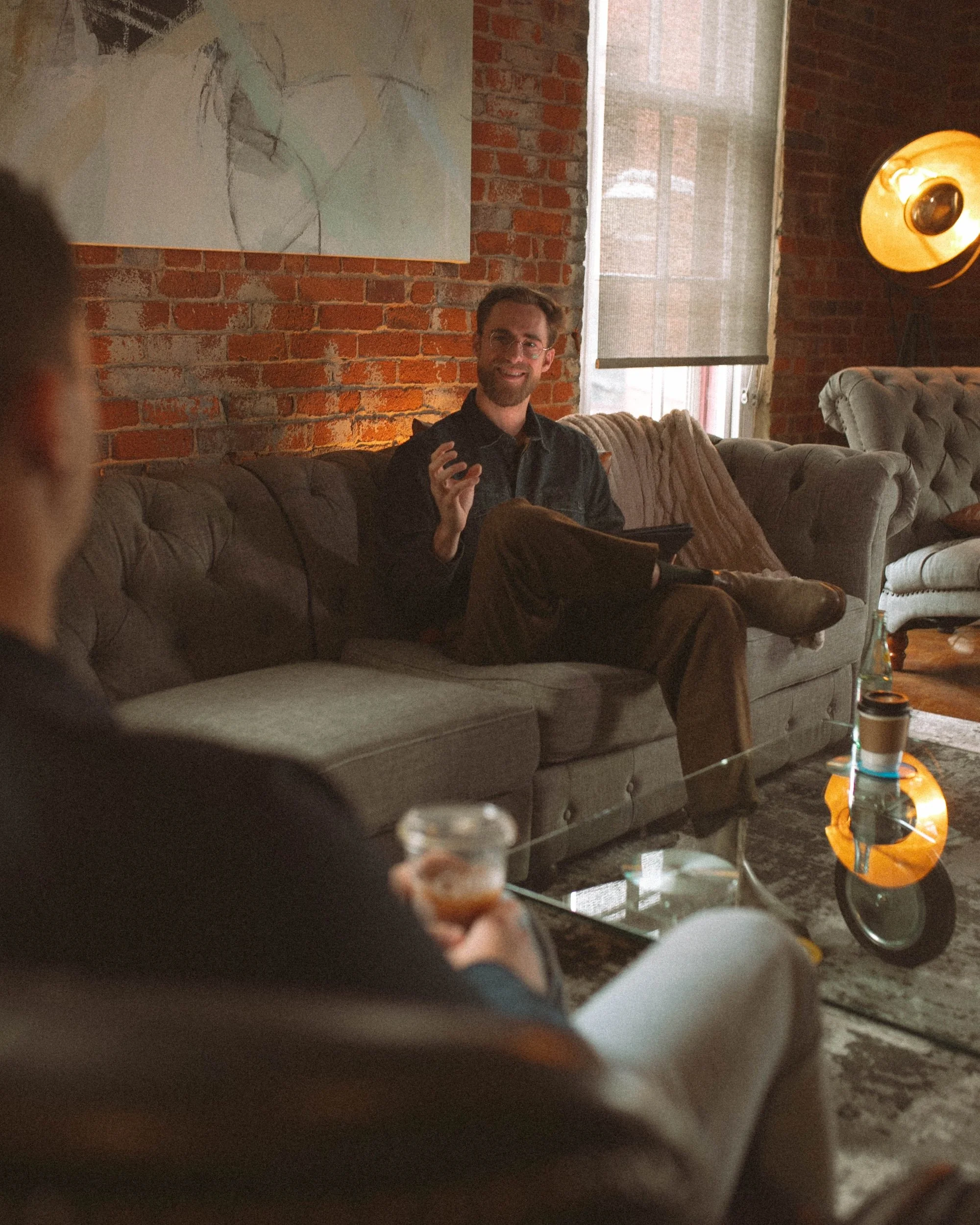 Brady Gilliam chats with a GWG faculty member on a tufted sofa in a sunlit brick loft.