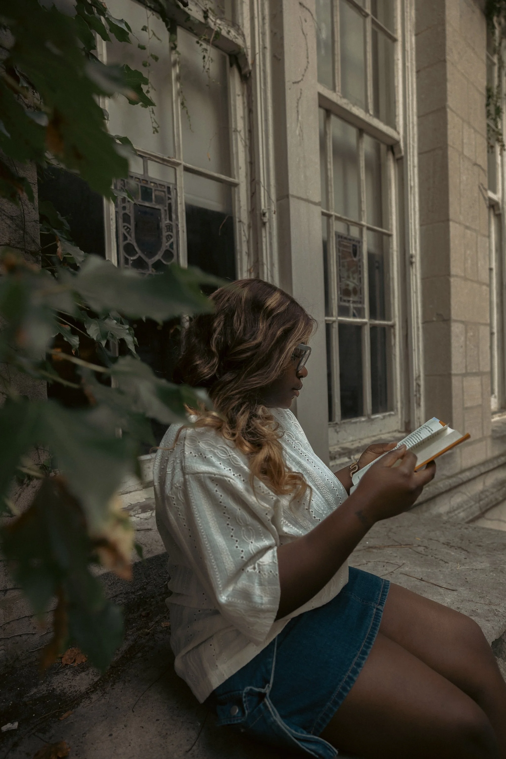 A GWG writing coach absorbed in a book sits outside an ivy-framed building's tall windows.