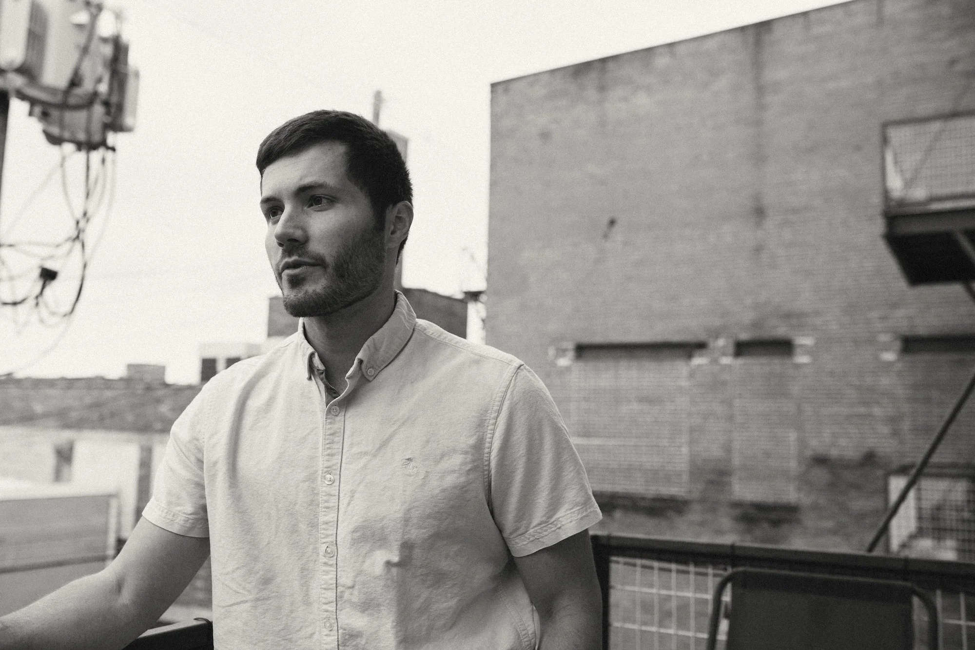 A writer in a light button-up shirt gazes off a rooftop near brick buildings.