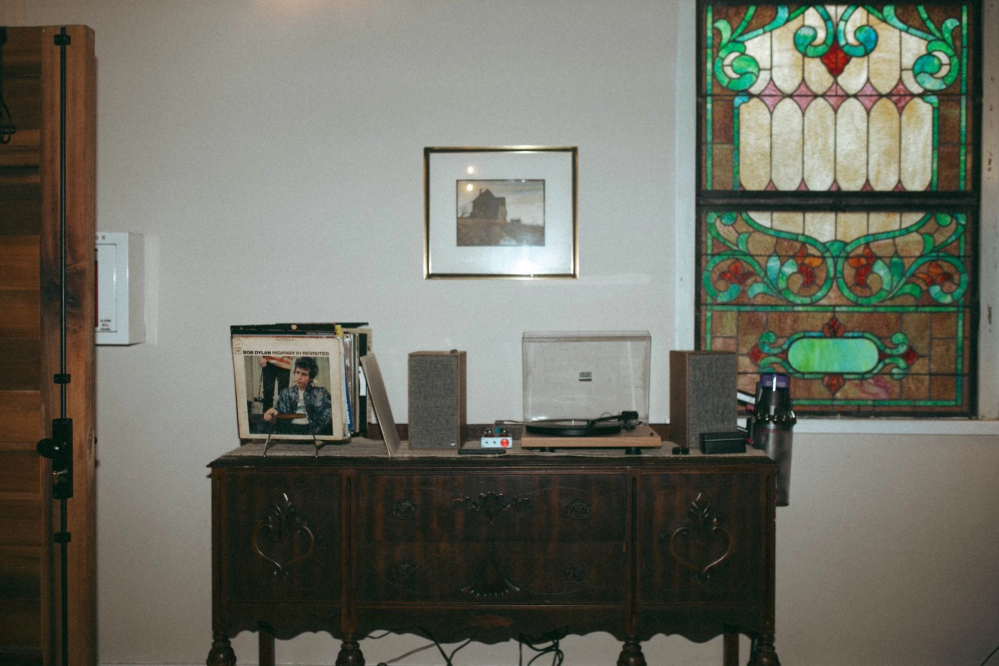 A vintage turntable and vinyl records on a credenza beside a stained-glass window.