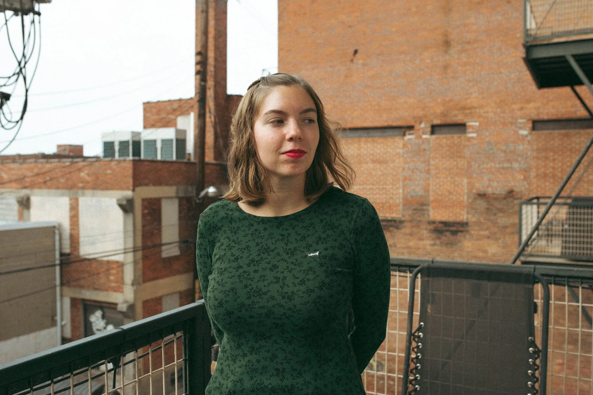 A GWG writing teacher in a green floral top stands on a balcony before brick warehouse walls.