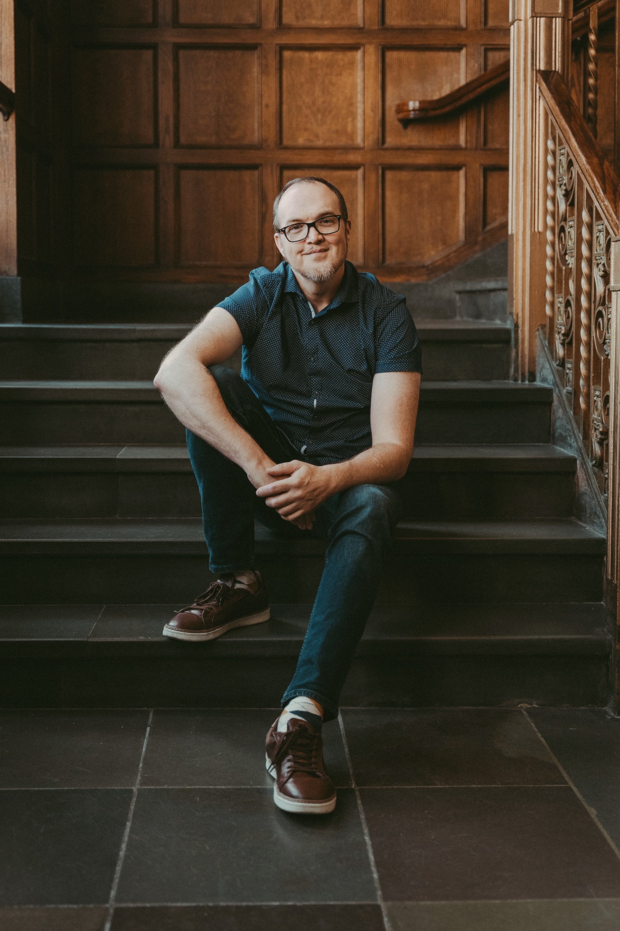 A bespectacled writing coach in a navy shirt sits smiling on a grand wood-paneled staircase.