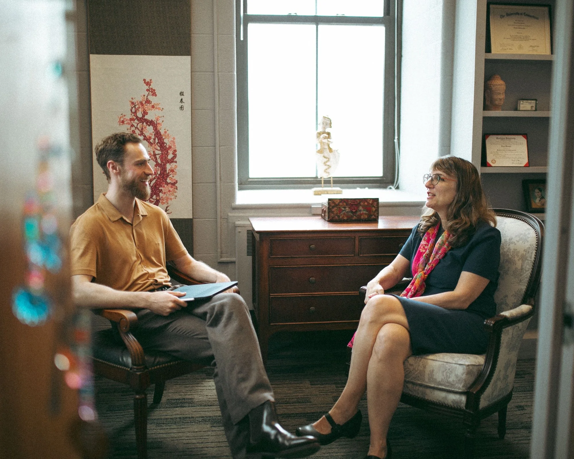 Brady Gilliam holds a notepad while interviewing a college professor in her diploma-lined office.