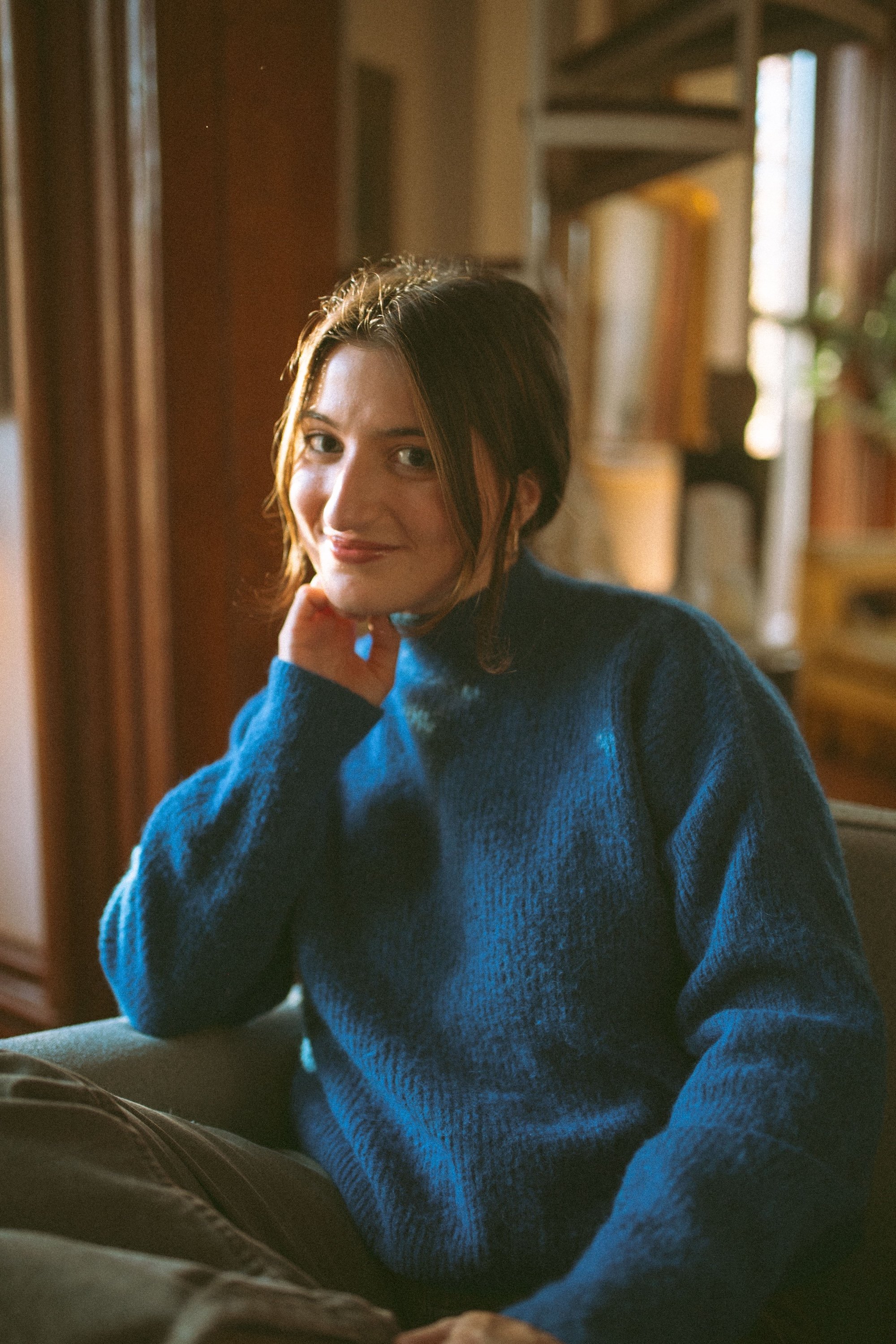 A writer in a blue knit sweater rests her chin against softly lit, book-filled shelves.
