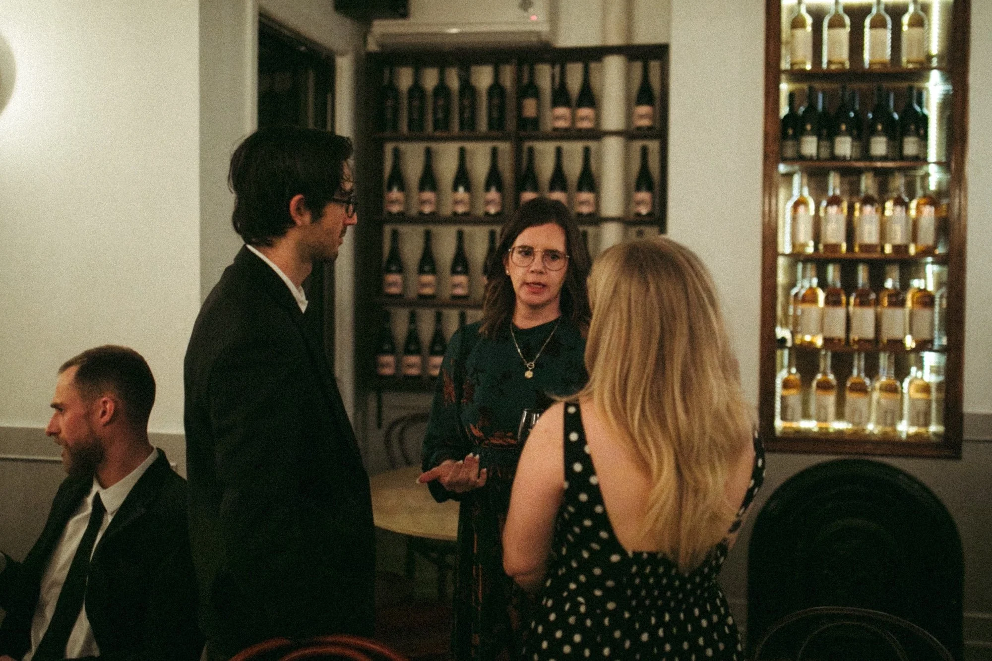Faculty talking over drinks beside a brightly lit wine display.