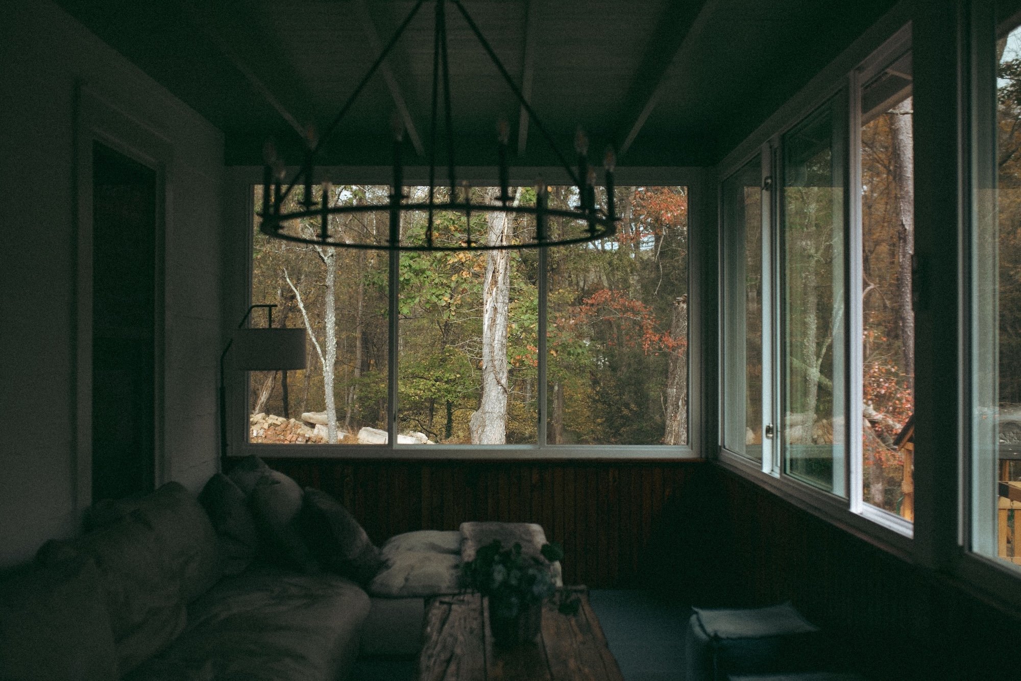A quiet sunroom with a chandelier and couch overlooking autumn woods.