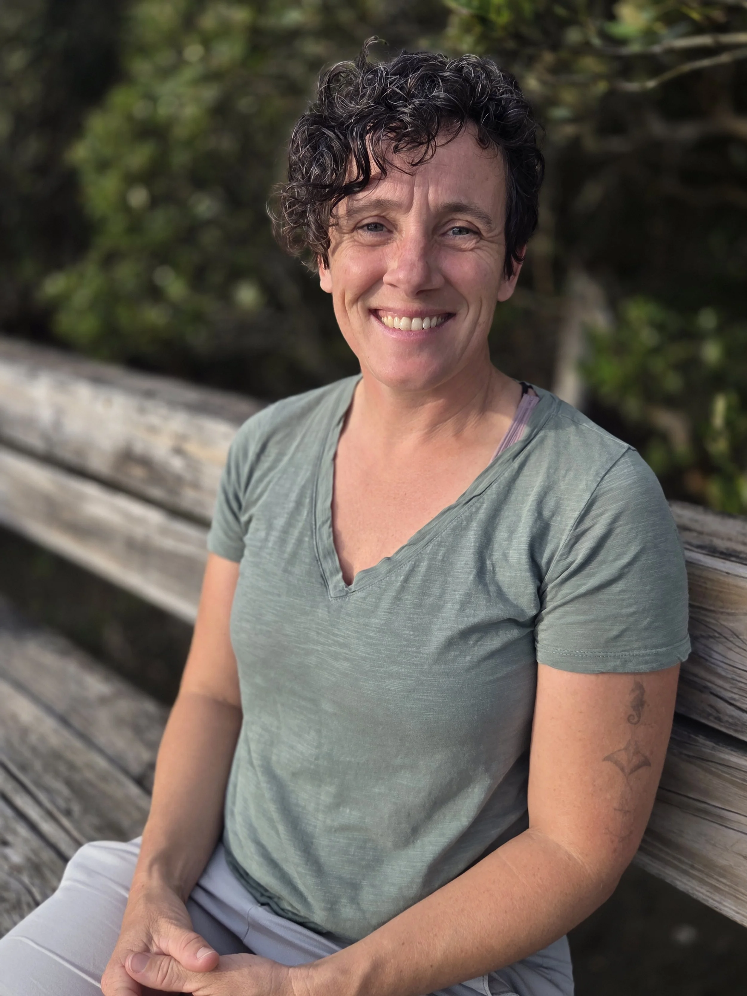 A woman with short, curly dark hair, smiling, wearing a green t-shirt, sitting on a wooden bench outdoors with a blurred green background.