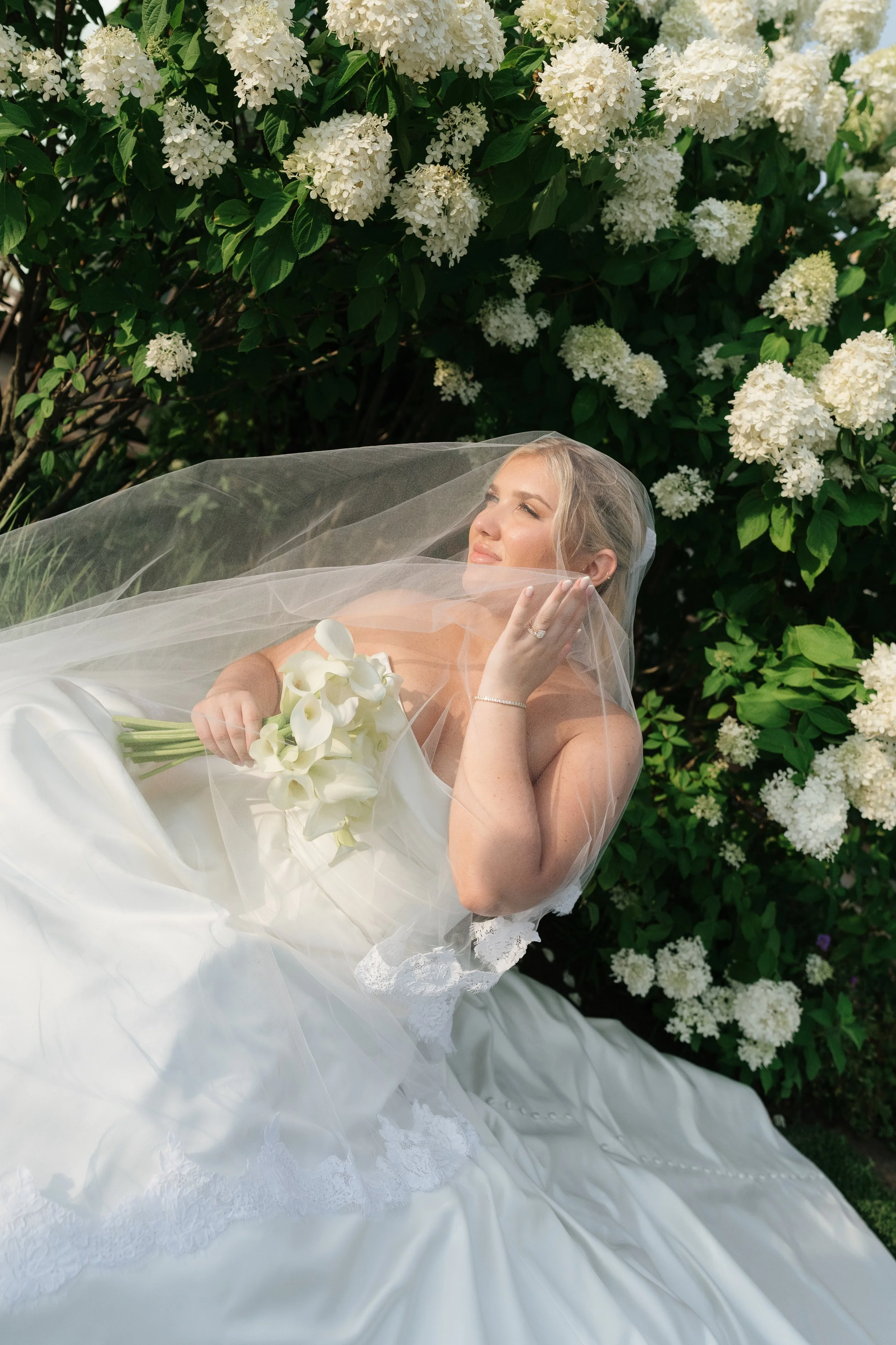 A woman in a wedding dress lying on the grass in front of a bush with white flowers, wearing a veil and holding a bouquet of white calla lilies.