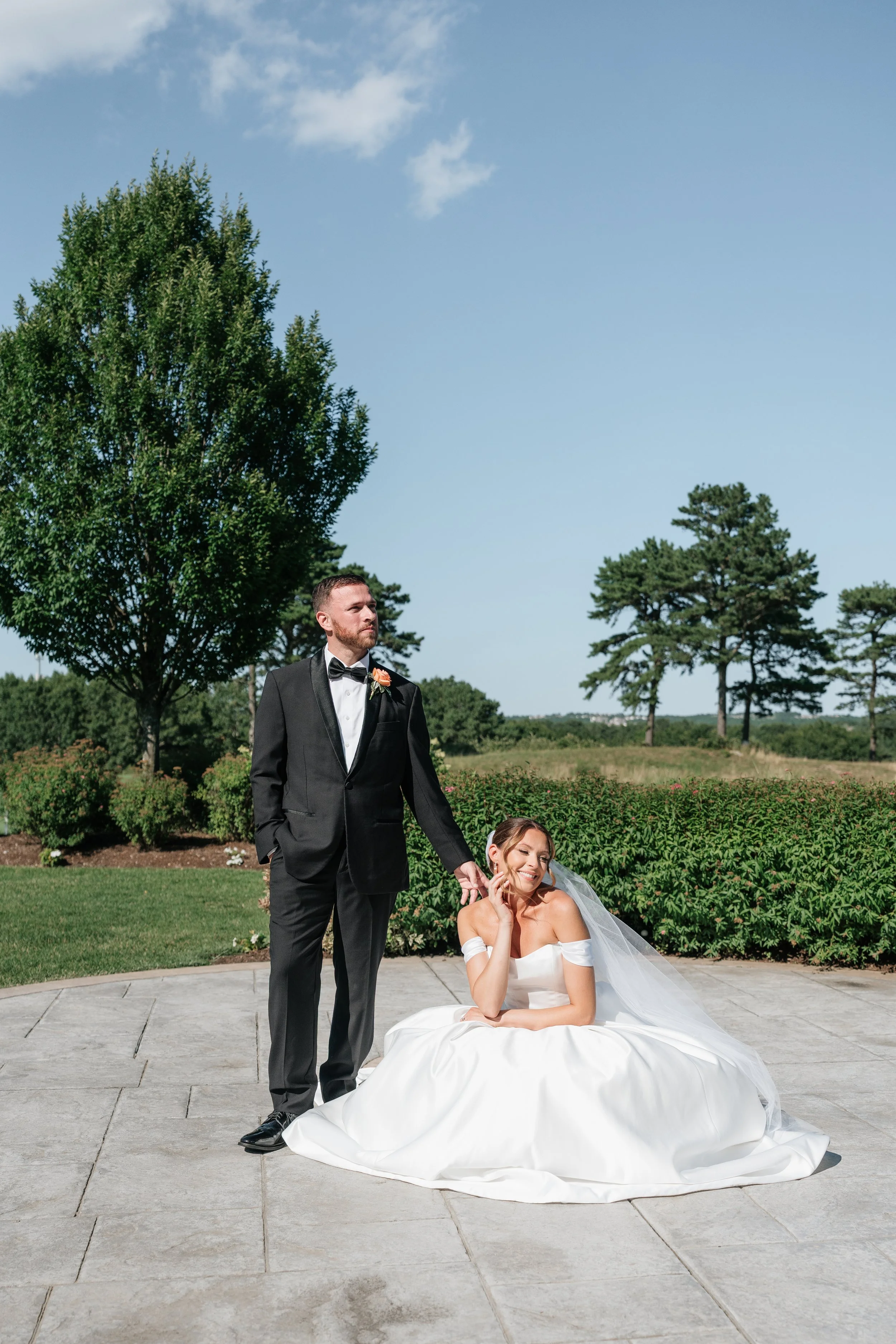 A bride sitting on the ground in a white wedding dress with her hand on her face, smiling, while a groom in a black tuxedo stands next to her holding her hand, outdoors on a patio with green trees and a blue sky in the background.
