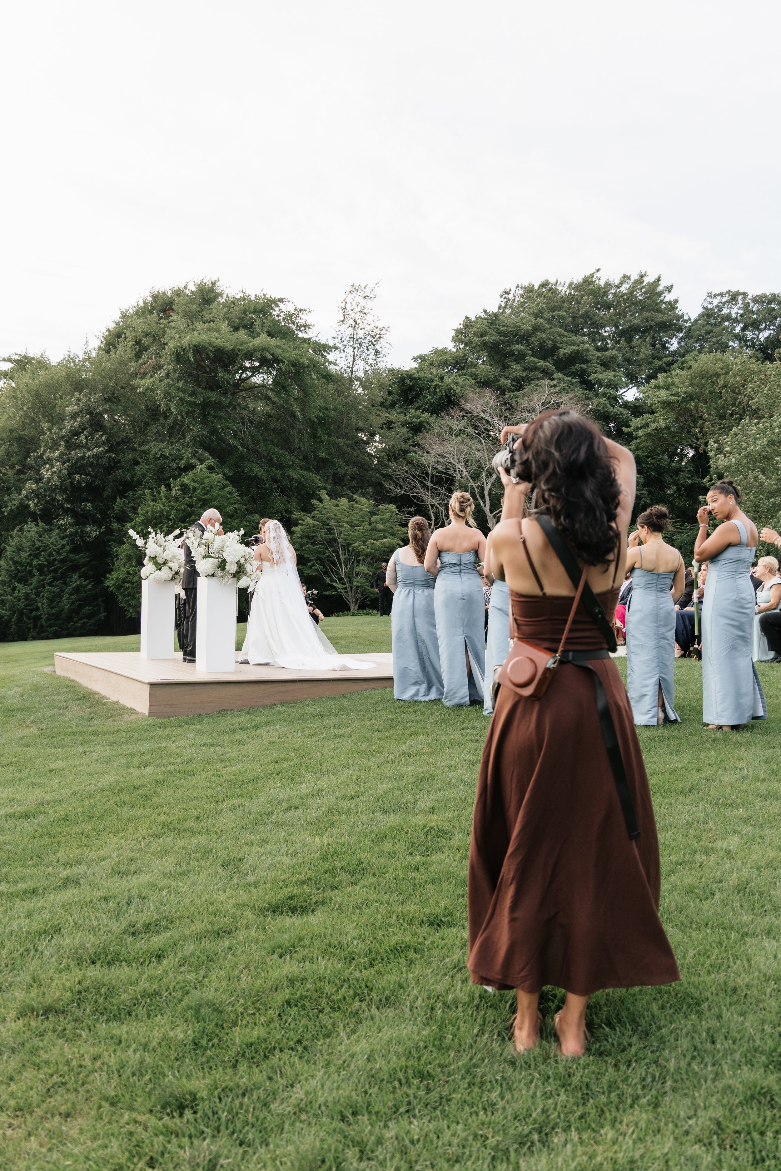 A woman dressed in a brown skirt and black top stands on a grassy lawn, taking a photograph of a wedding ceremony happening in front of her with a camera. The ceremony involves a bride and groom at an altar decorated with white flowers, and a group of bridesmaids in light blue dresses observing.