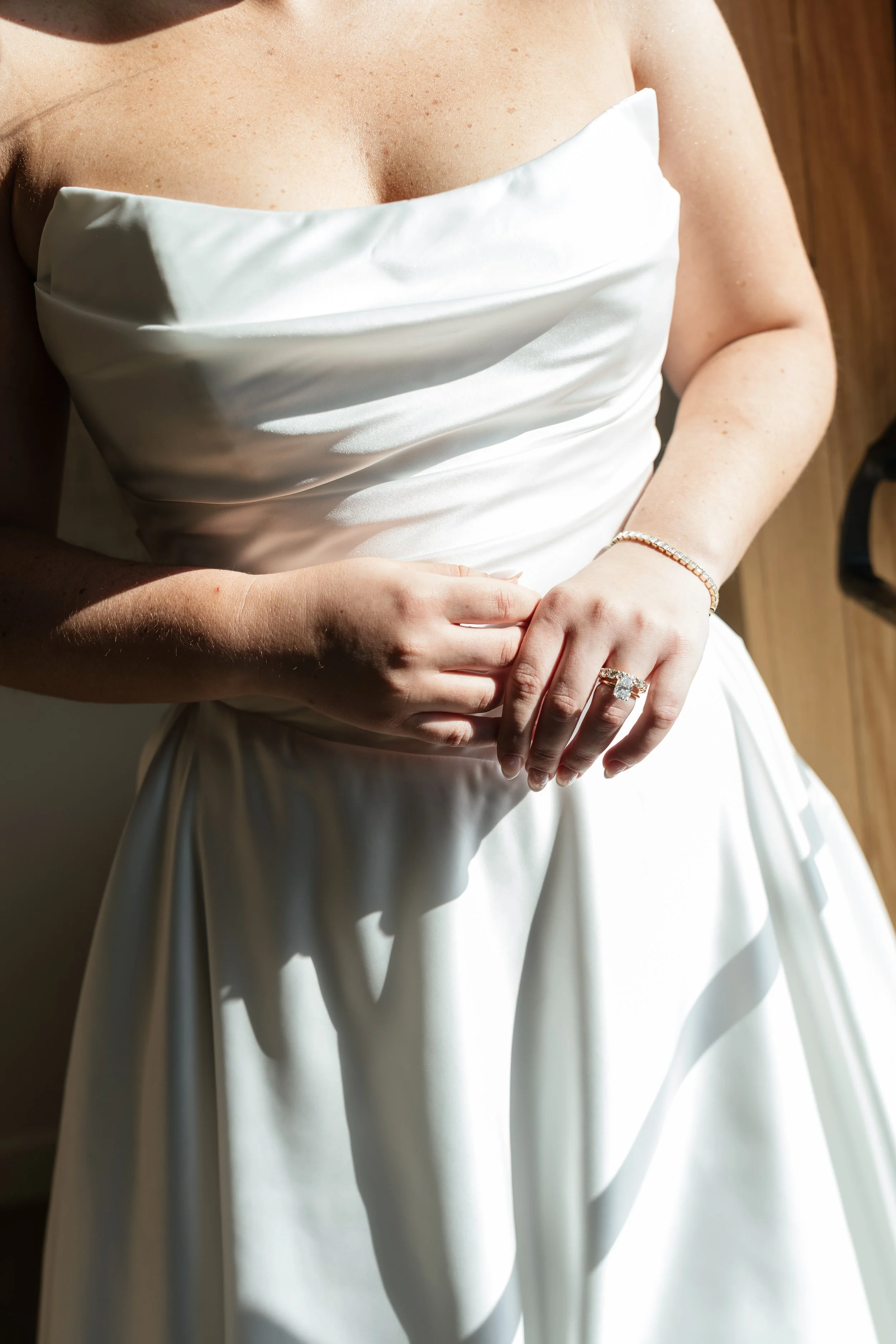 Close-up of a woman in a white satin dress, showing her upper torso and hand with a diamond ring and bracelet, against a wooden background.