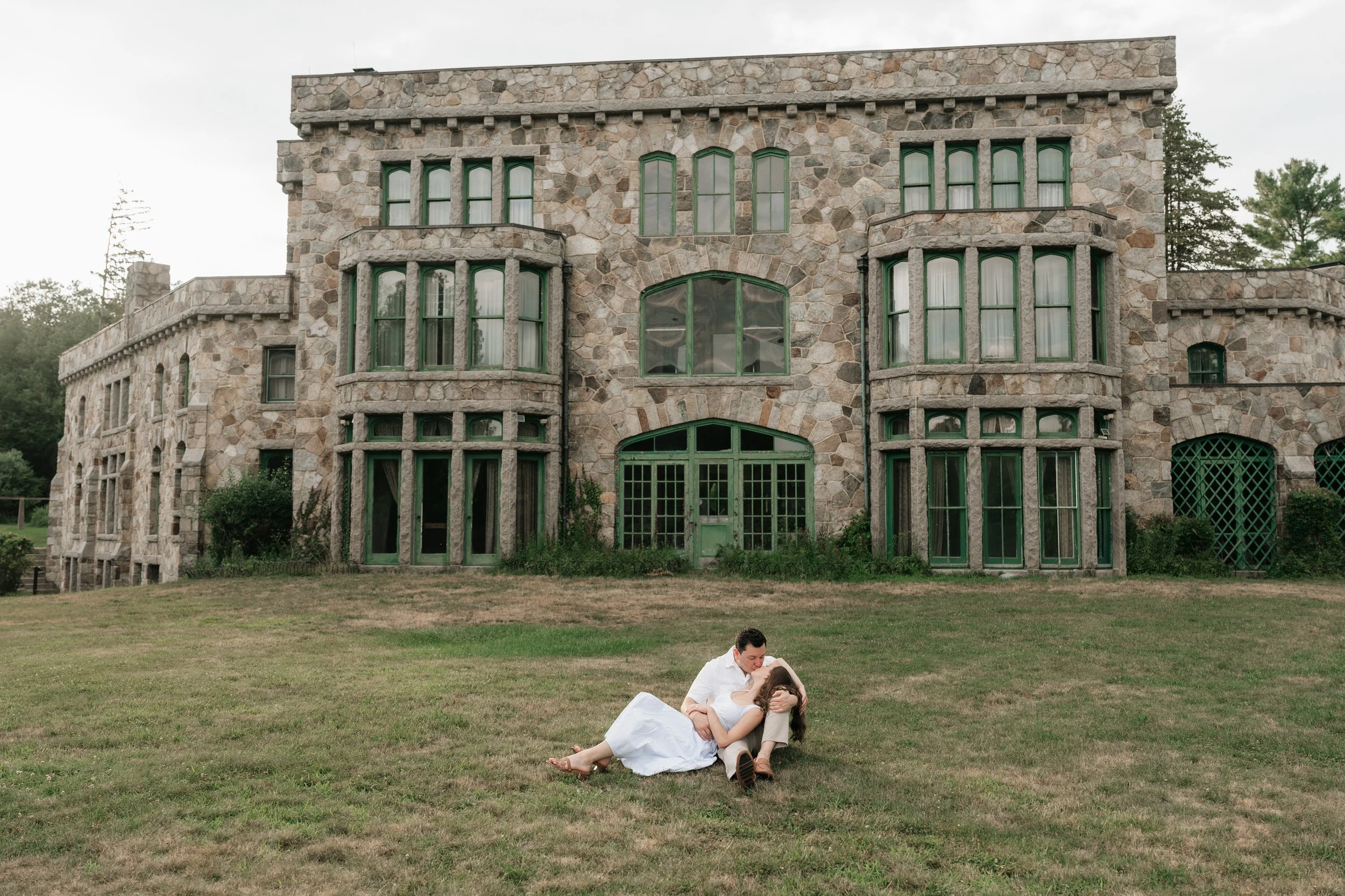 A man and woman embracing on a grassy lawn in front of a large stone building, with the woman lying on the man’s lap. The man is wearing a white shirt and tan pants, and the woman is in a white dress.