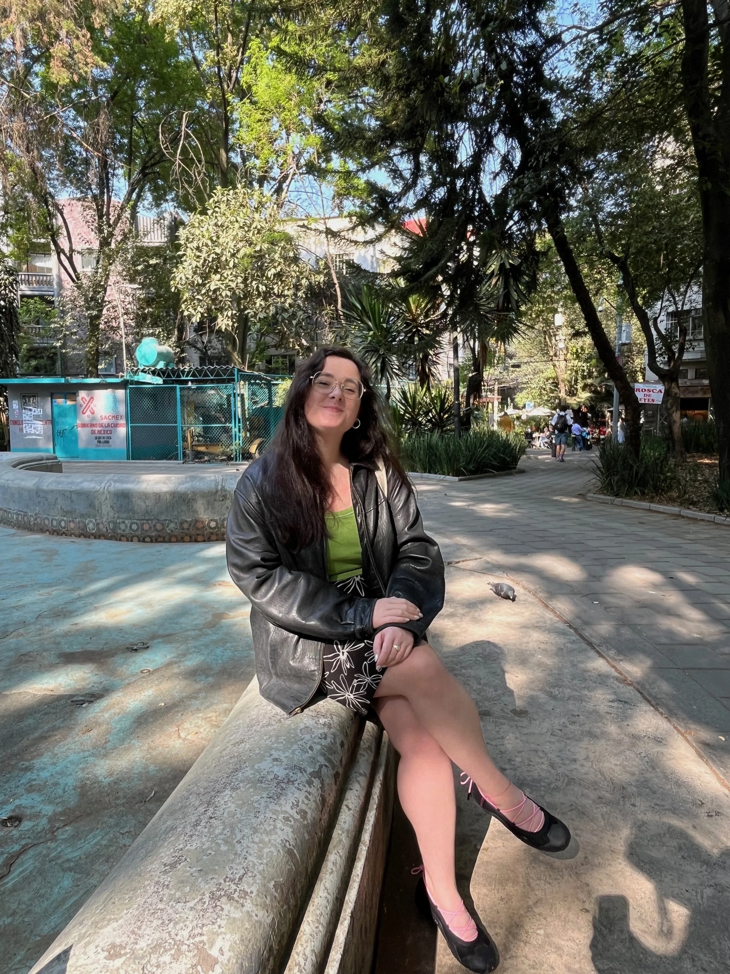 A woman is sitting on a stone bench in a park, surrounded by trees and greenery. She is wearing glasses, a black leather jacket, a green top, and a black skirt with white floral patterns. She has pink ballet flats with ribbons. The scene includes a fountain, greenery, and a street with pedestrians and buildings in the background.