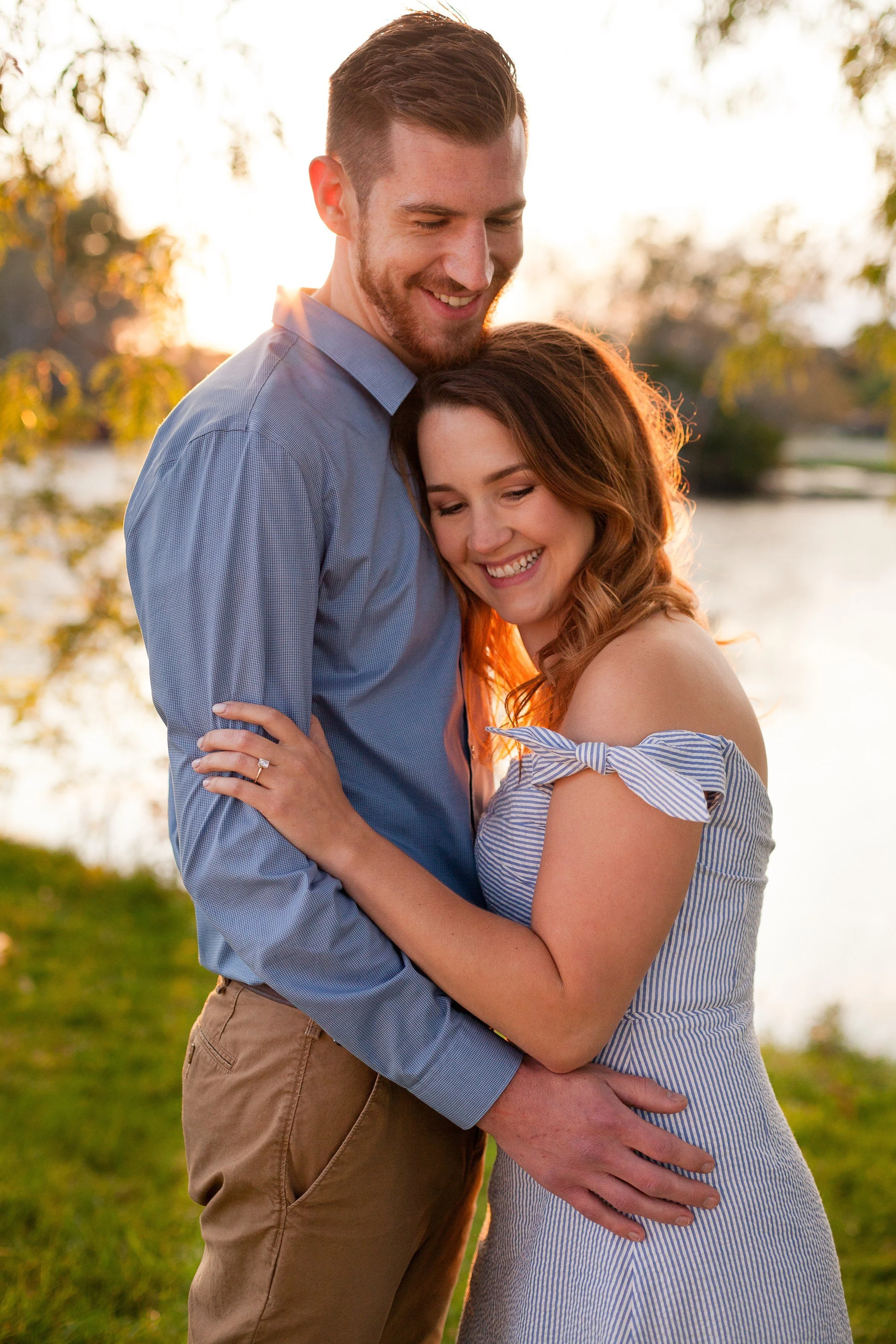 A couple hugging outdoors near a body of water at sunset, smiling and close together.