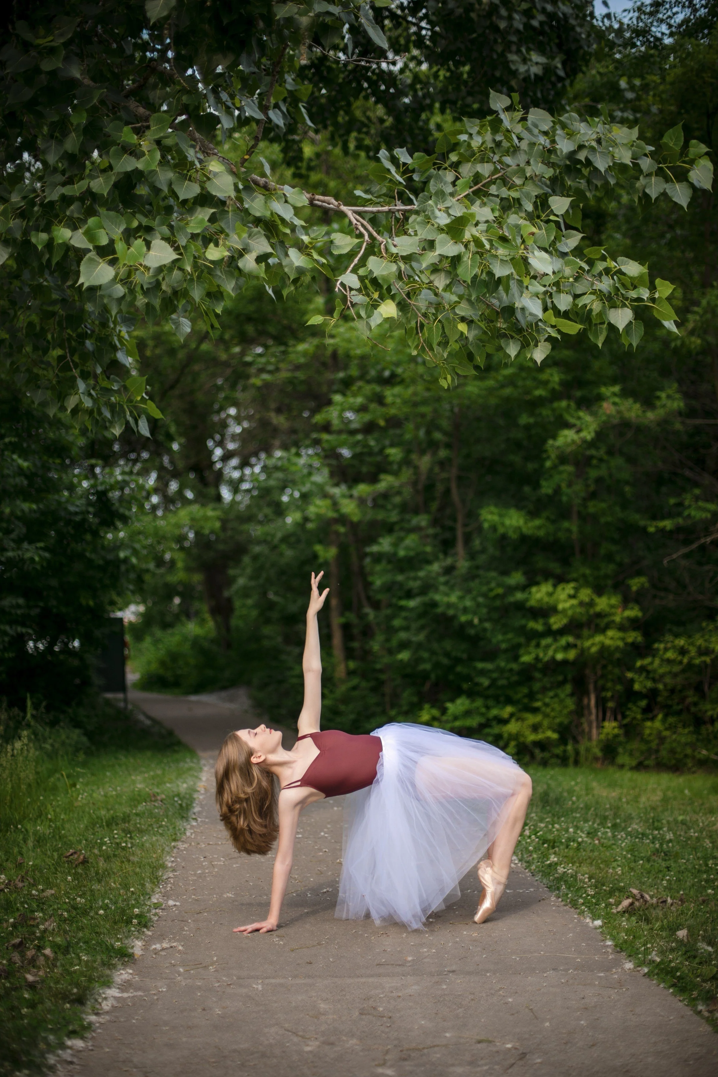 A ballerina in a burgundy top and white tulle skirt performs a pose on an outdoor path surrounded by green trees, reaching upward with one arm and balancing on one foot.