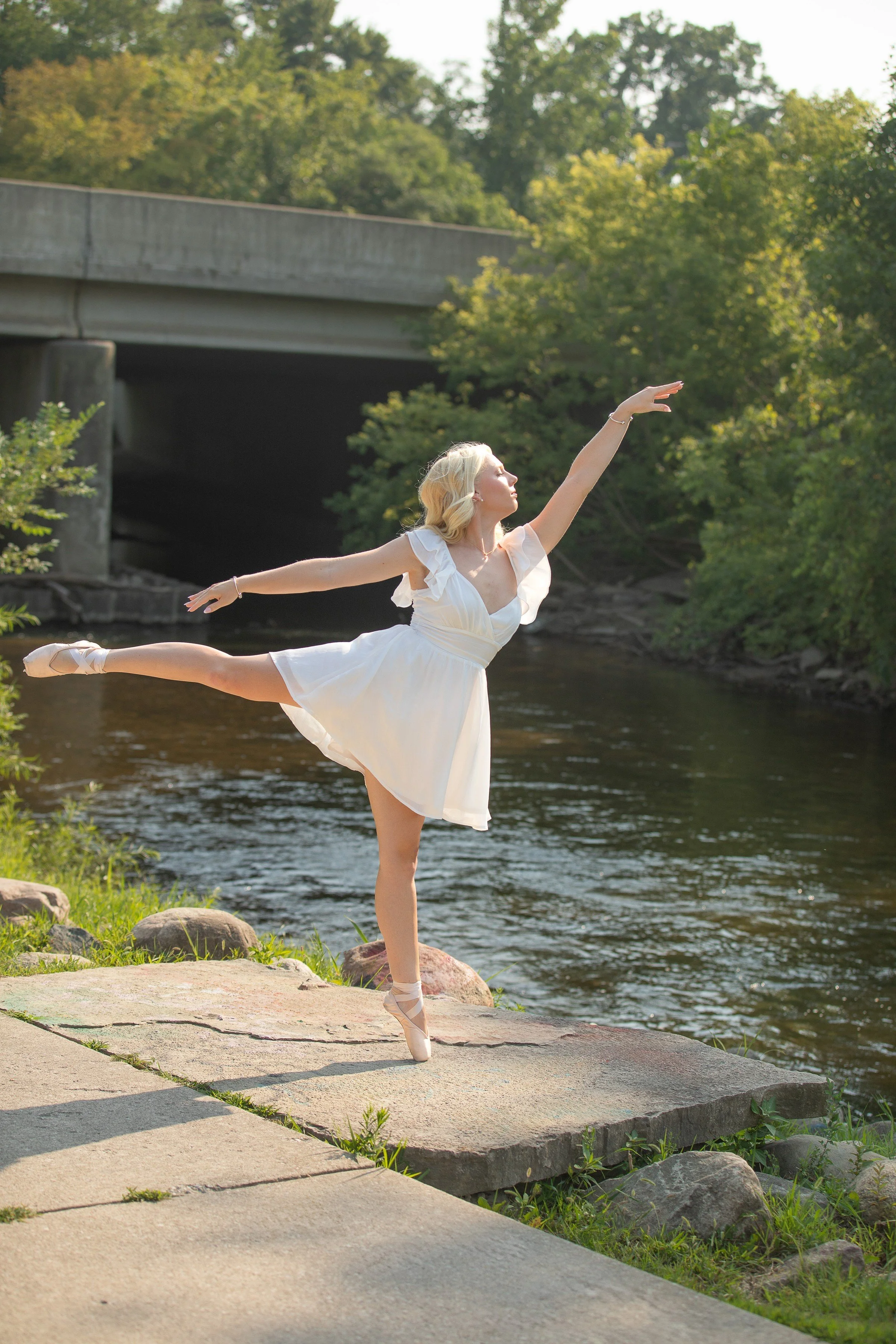 A woman in a white dress is ballet dancing outdoors by a river, standing on one foot with the other leg extended behind and arms raised gracefully.