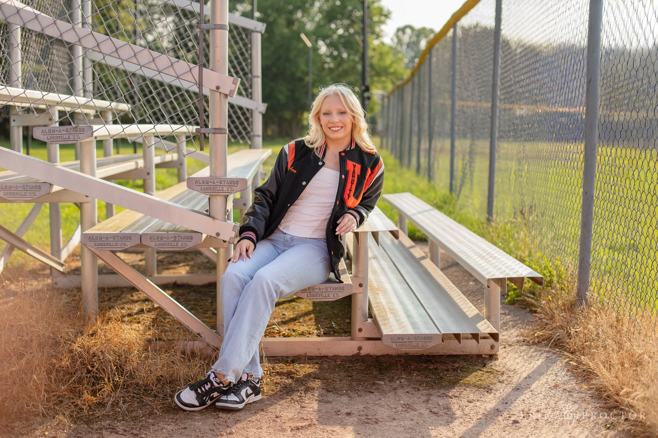 A young woman sitting on metal bleachers at an outdoor sports field, wearing a black and red varsity jacket, light gray sweatpants, and black and white sneakers, with a chain necklace, smiling in the sunlight.