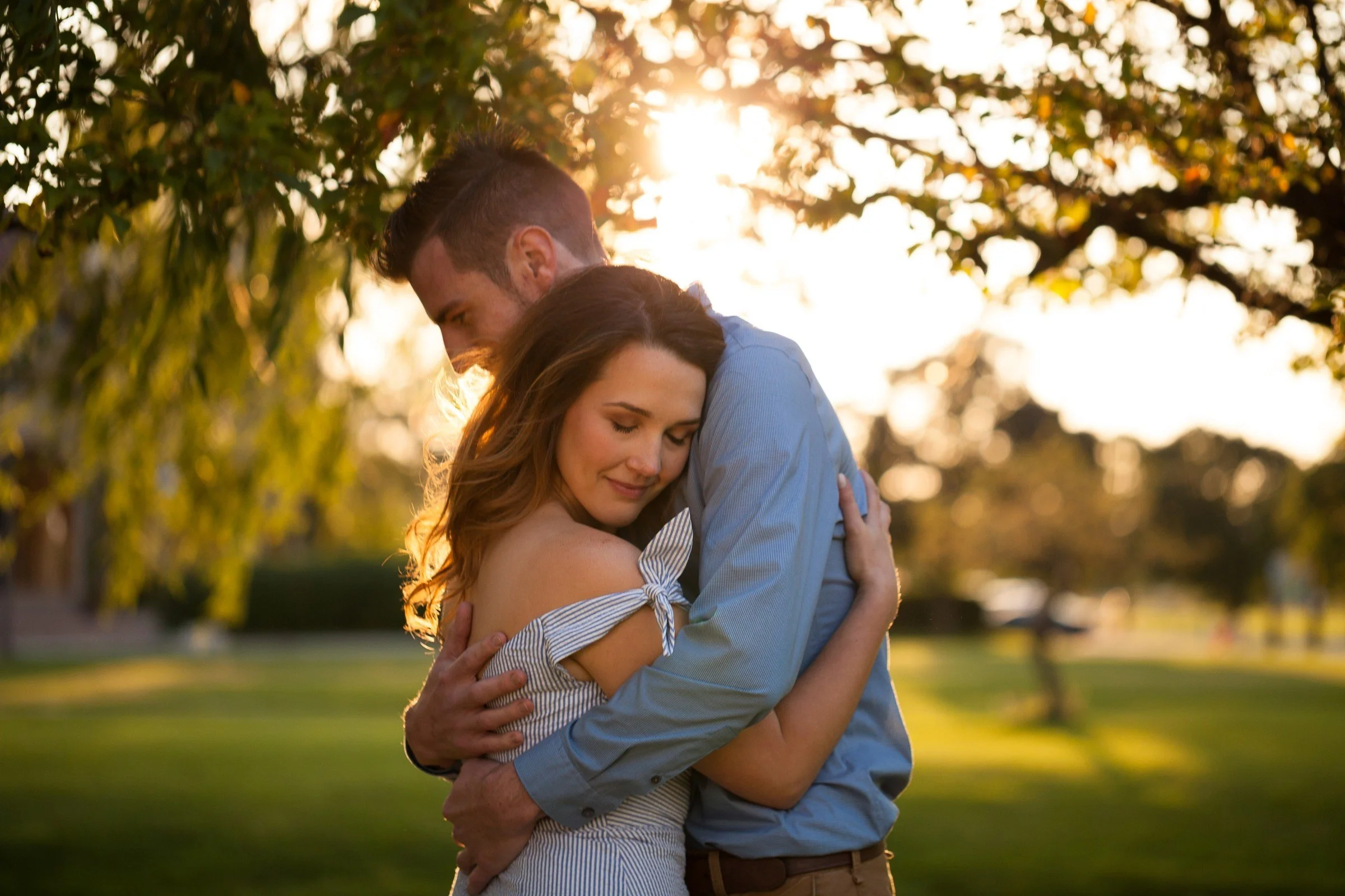 A couple hugging in a park during sunset. The woman has long brown hair and is wearing a striped dress with off-shoulder sleeves. The man has short hair and a beard, wearing a blue shirt. They are embracing with closed eyes, and trees with green leav