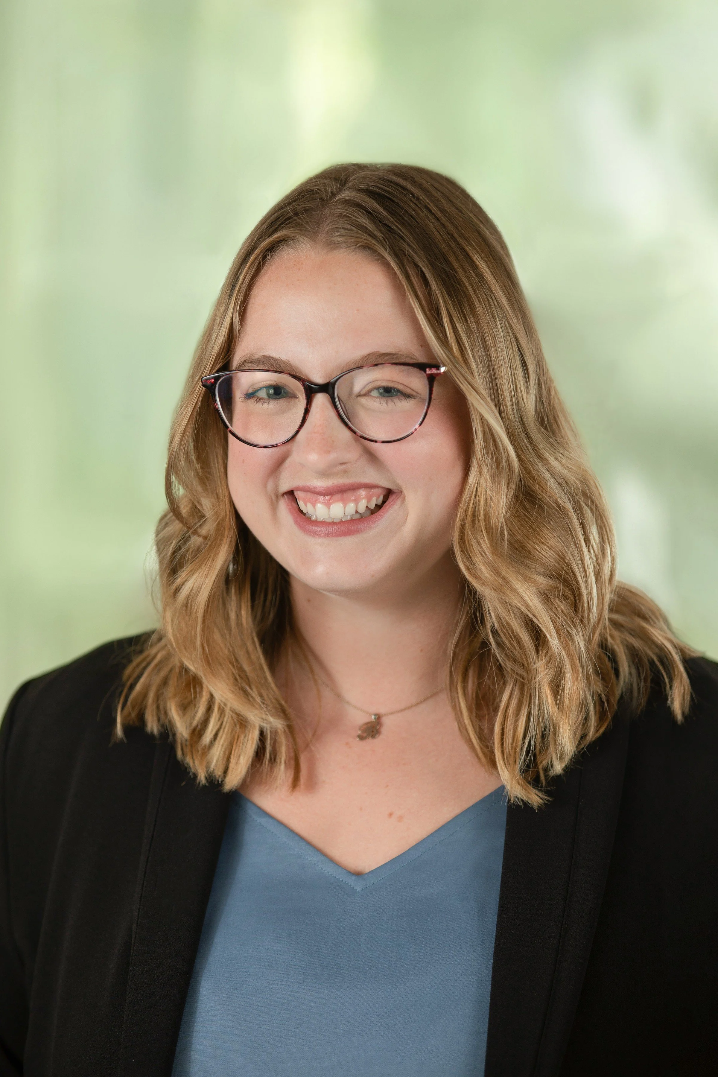 A young woman with shoulder-length wavy blonde hair, black-rimmed glasses, and a bright smile, wearing a dark blazer over a blue top, standing against a blurred green background.