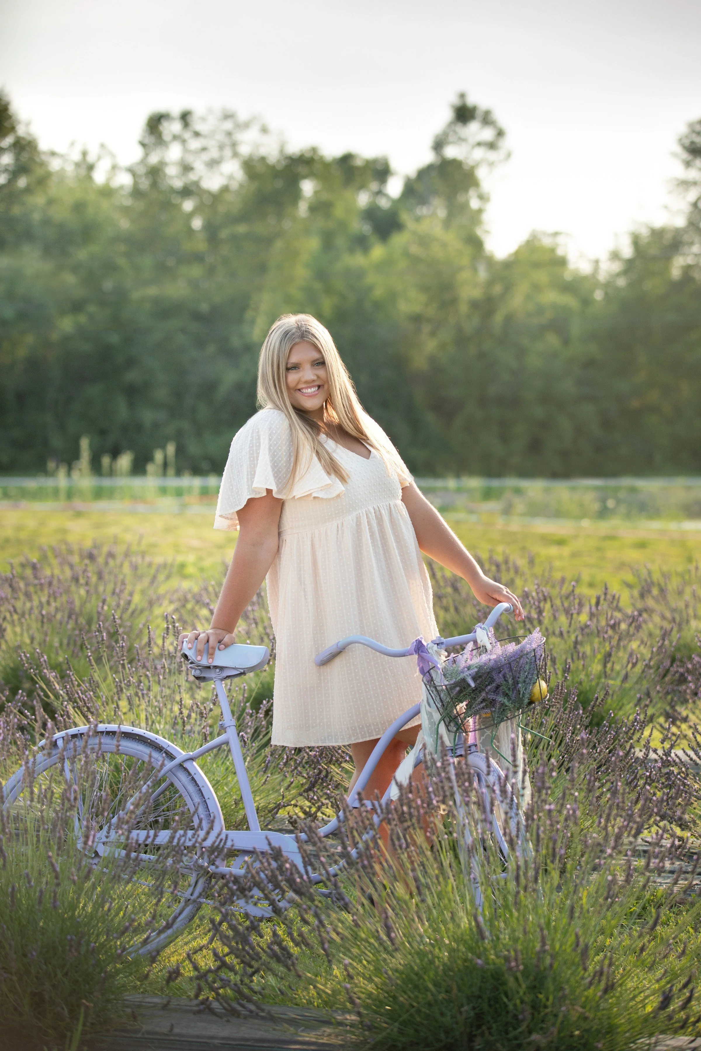 A smiling woman in a light yellow dress standing outdoors with a purple bicycle in a lavender field, with trees in the background.