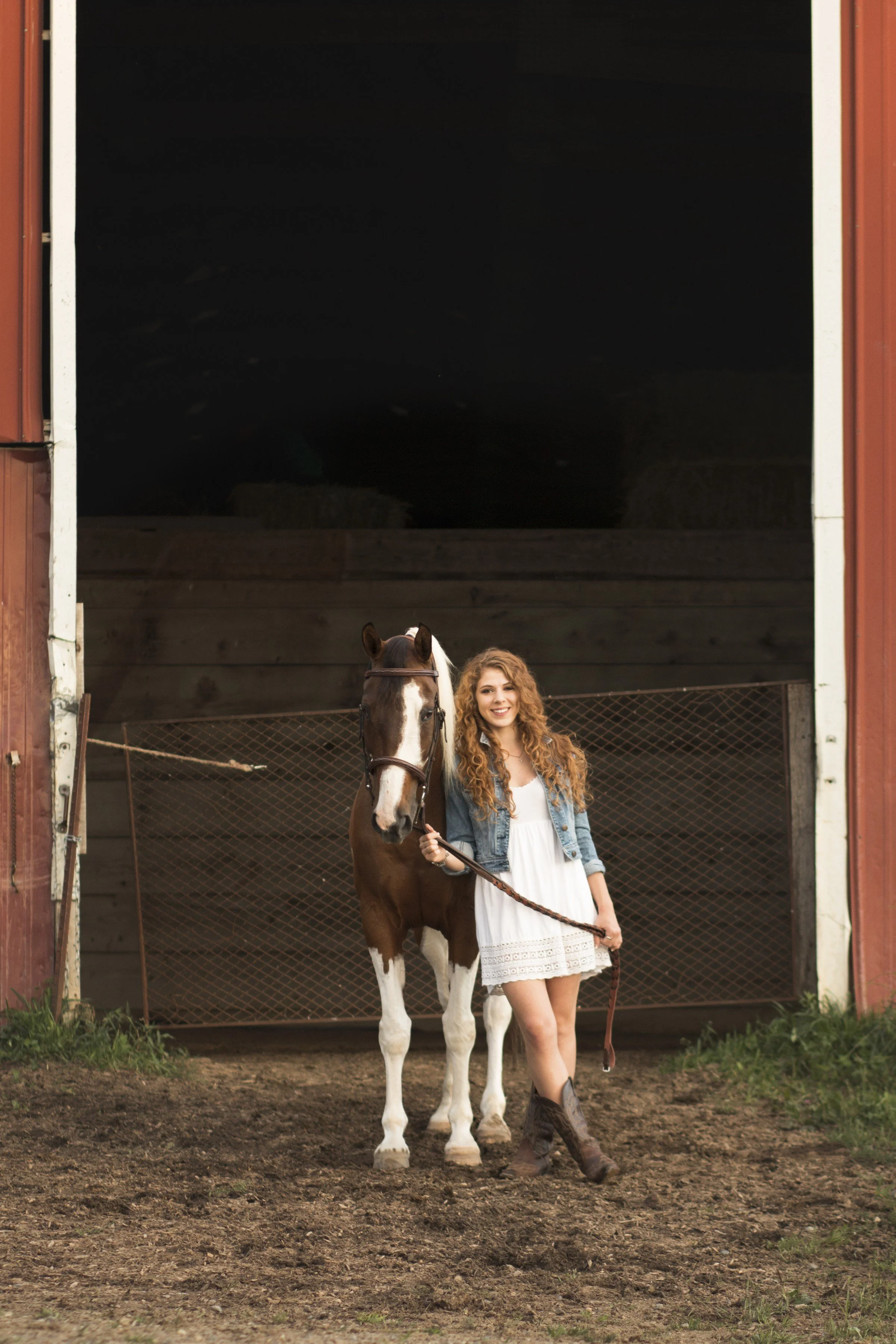 A young woman with curly red hair wearing a white dress, denim jacket, and cowboy boots standing next to a brown and white horse inside a barn.