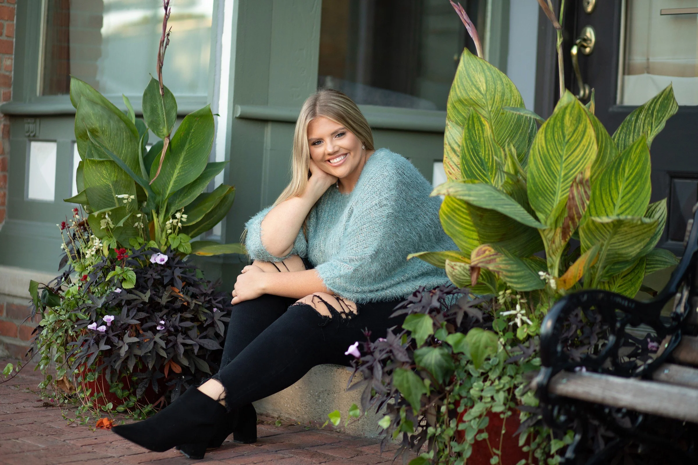 A young woman with blonde hair sitting on the front porch of a house, smiling, surrounded by large green and purple potted plants.