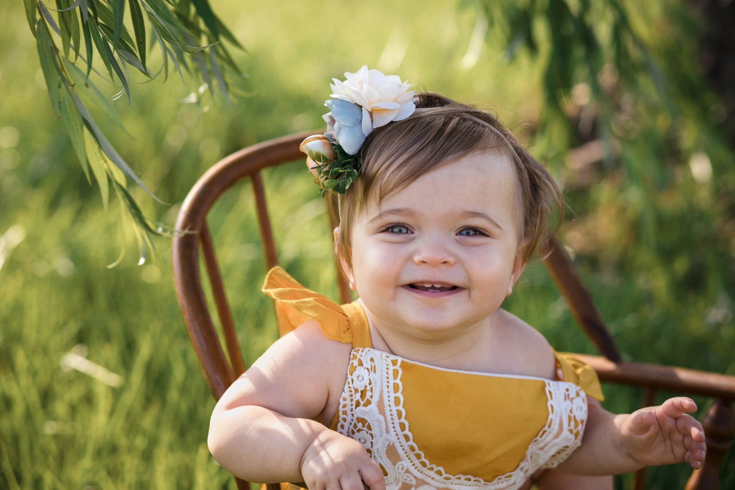 A smiling baby girl with light blue eyes, wearing a yellow dress with white lace detail and floral shoulder accents, sitting in a wooden chair outdoors amid lush green foliage, with a large white flower headband.