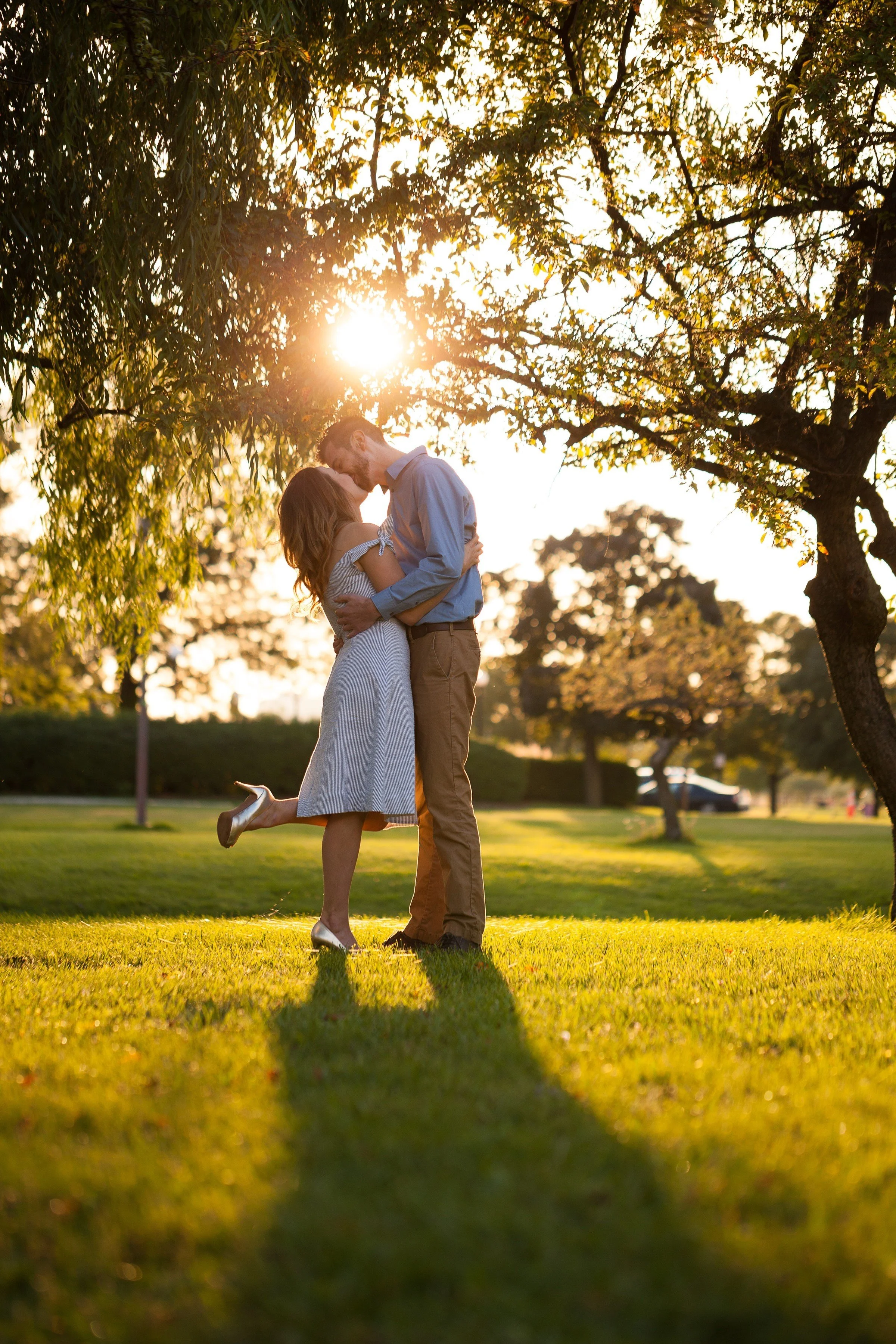 A couple kissing outdoors at sunset, with the woman lifting one foot in the air, standing on grass under trees.