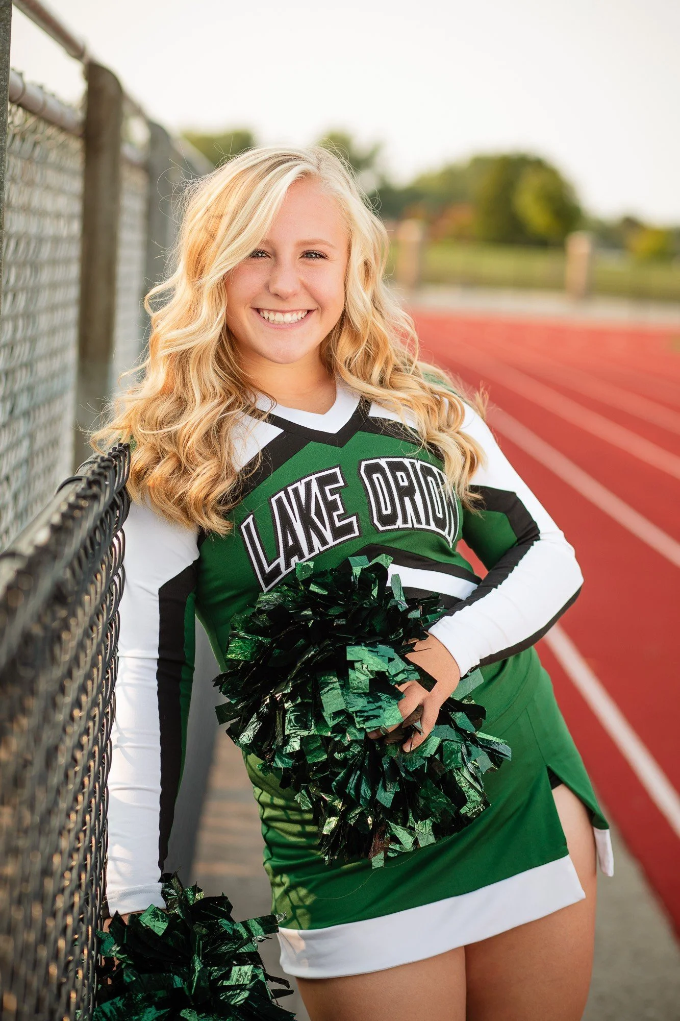 A young female cheerleader with blonde curly hair, smiling, wearing a green and white cheerleading uniform with 'Lake Orion' on it, holding green and black pom-poms, standing next to a chain-link fence on a track field.