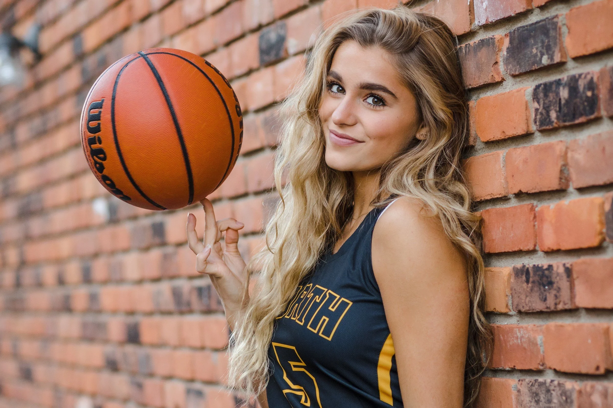 Young woman in a basketball jersey leaning against a brick wall holding a basketball on one finger, smiling at the camera.
