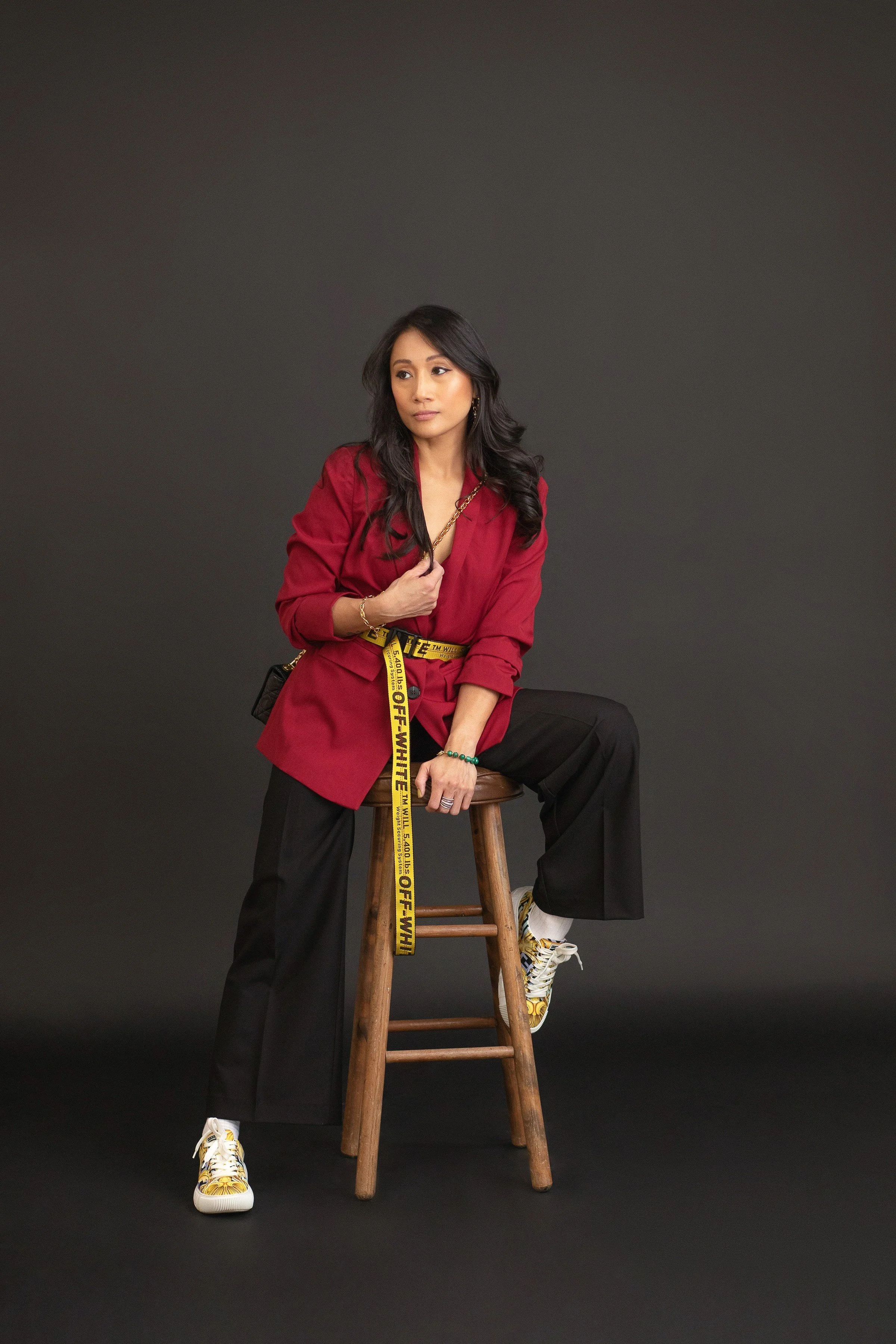 A woman with long black hair sitting on a wooden stool against a dark background, wearing a red blazer, black wide-leg pants, and yellow and white shoes, with a yellow Off-White belt.