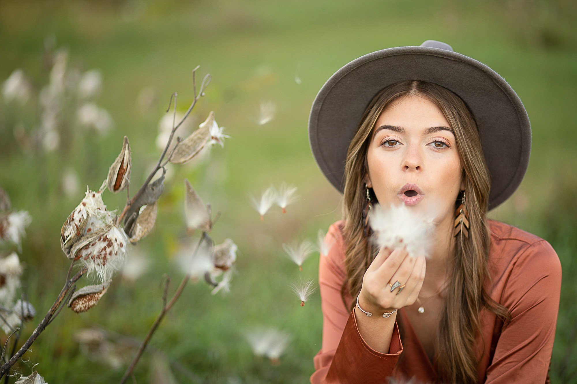 Woman with long hair, wearing a gray hat and earrings, blowing on cottonwood fluff in a green outdoor setting.