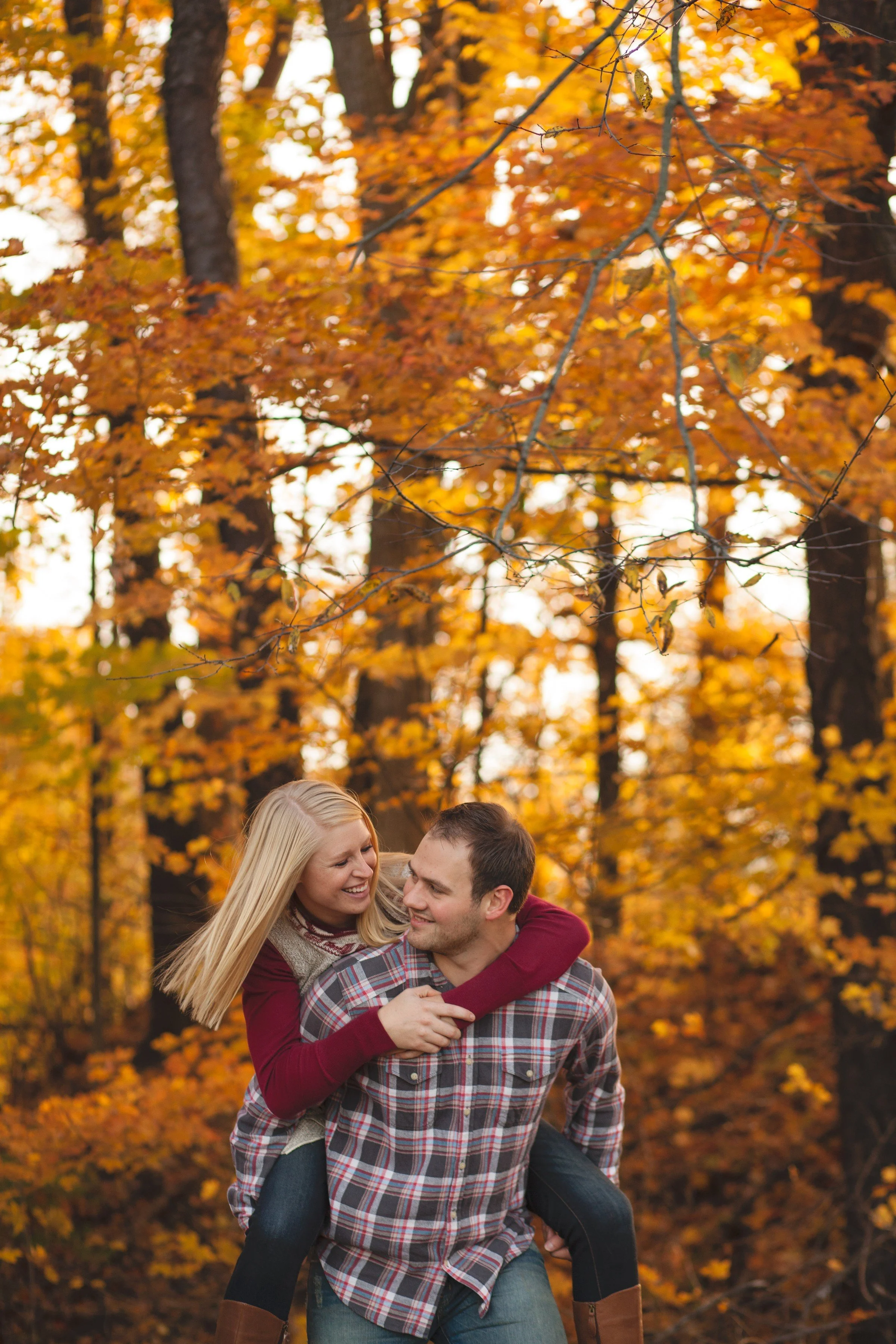 A smiling couple, a woman on a man's back, in a forest with autumn leaves.