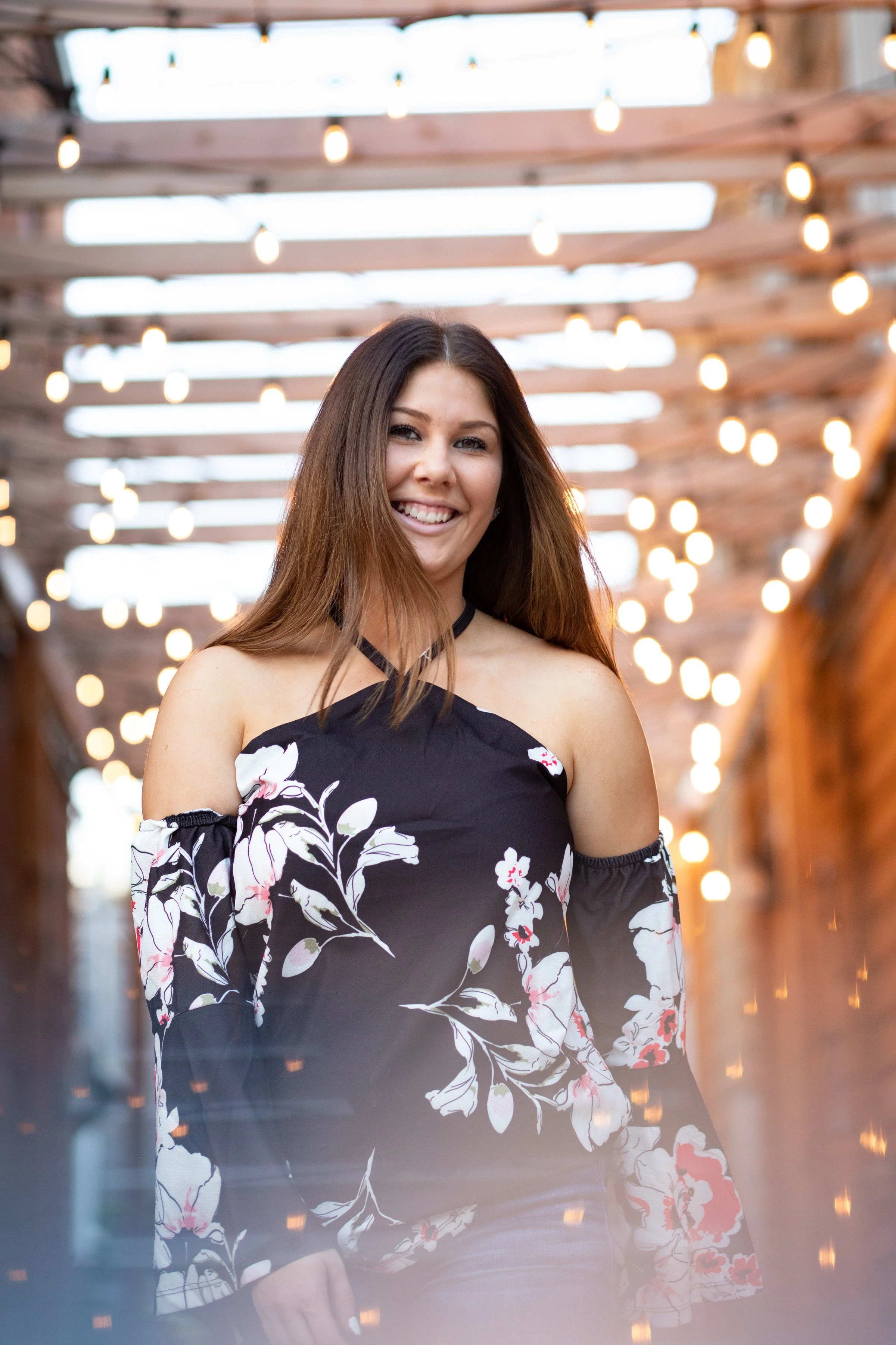 A young woman with long brown hair smiling, wearing a black floral off-the-shoulder top, standing underneath a wooden pergola decorated with string lights.
