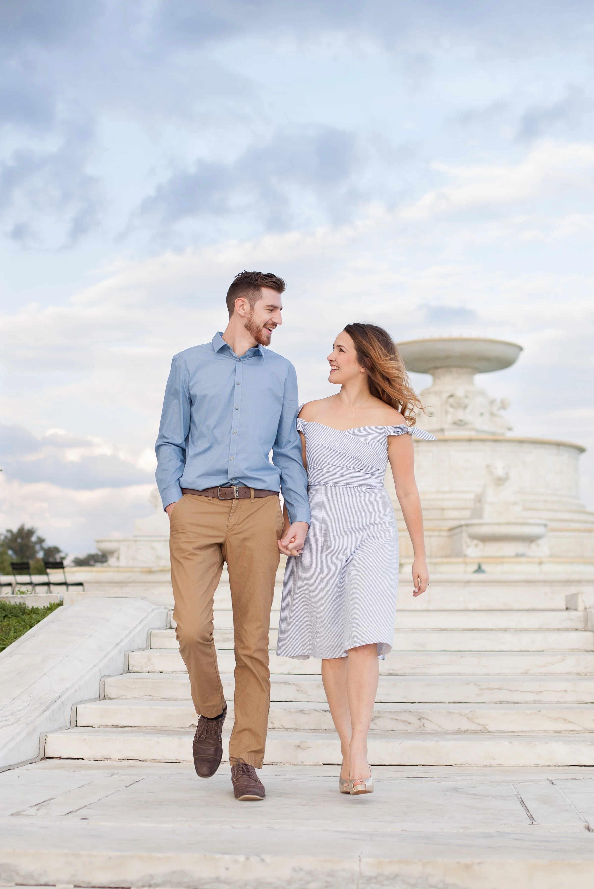 A couple walking hand-in-hand outdoors near a white marble fountain, smiling at each other against a partly cloudy sky.