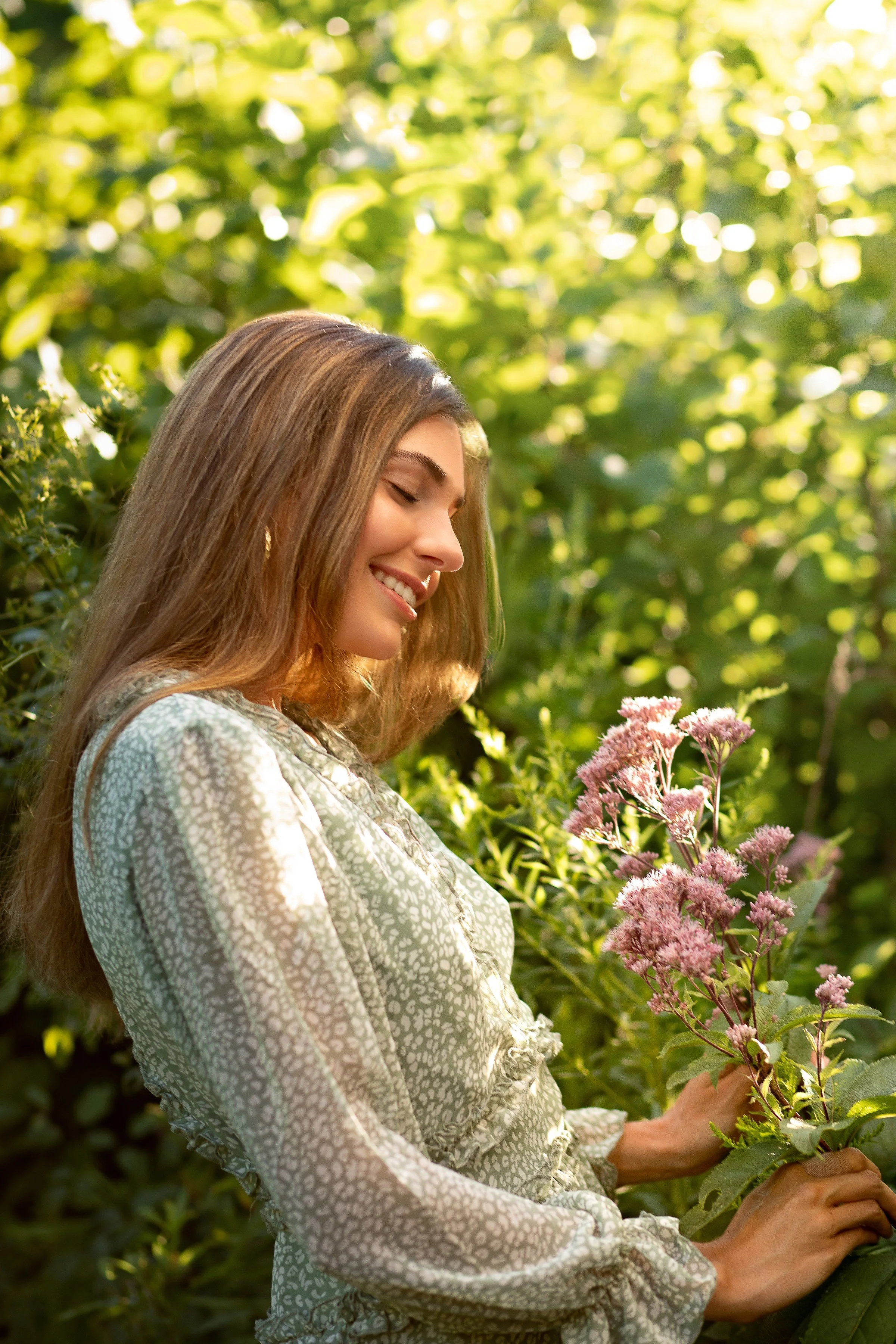 A woman with long, light brown hair smiling and holding a bunch of pink flowers in a green, sunlit outdoor setting.