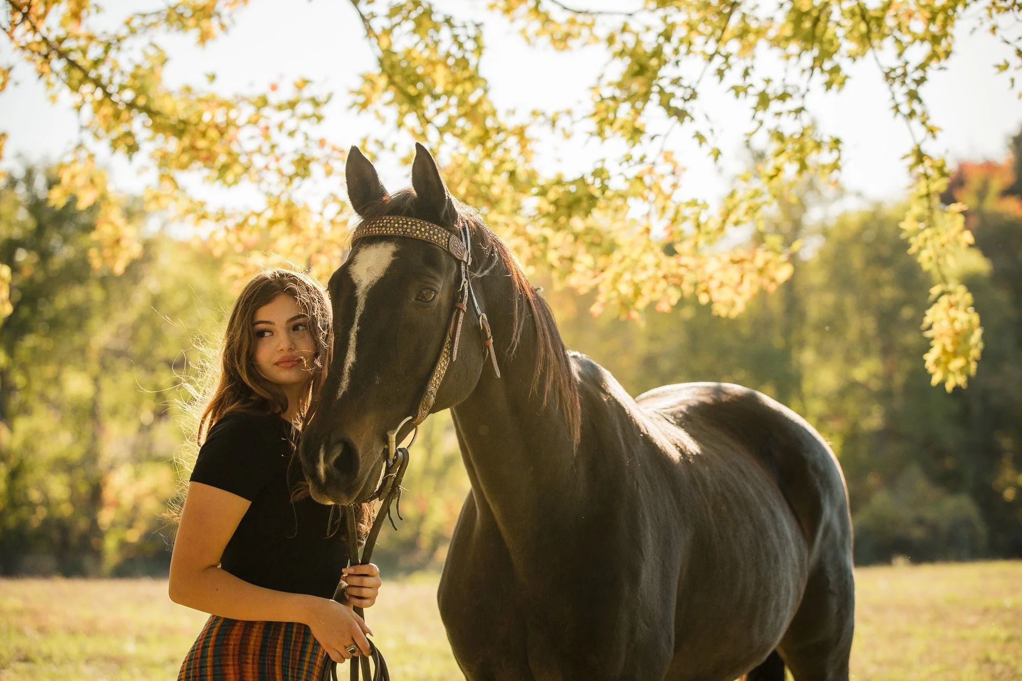 A young woman standing next to a black horse with a white blaze on its face outdoors during fall, with trees and leaves in the background.