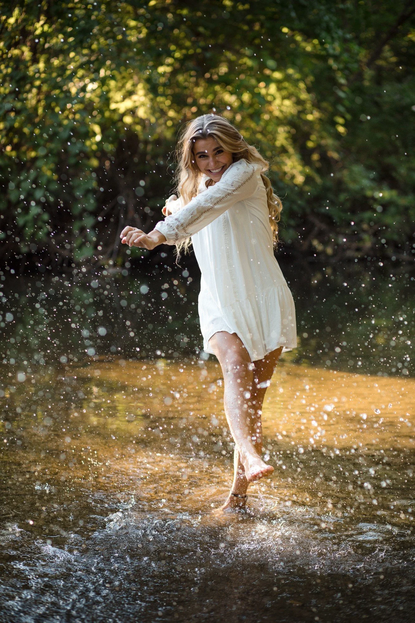 A woman in a white dress joyful playing and splashing water in a shallow creek surrounded by green trees.