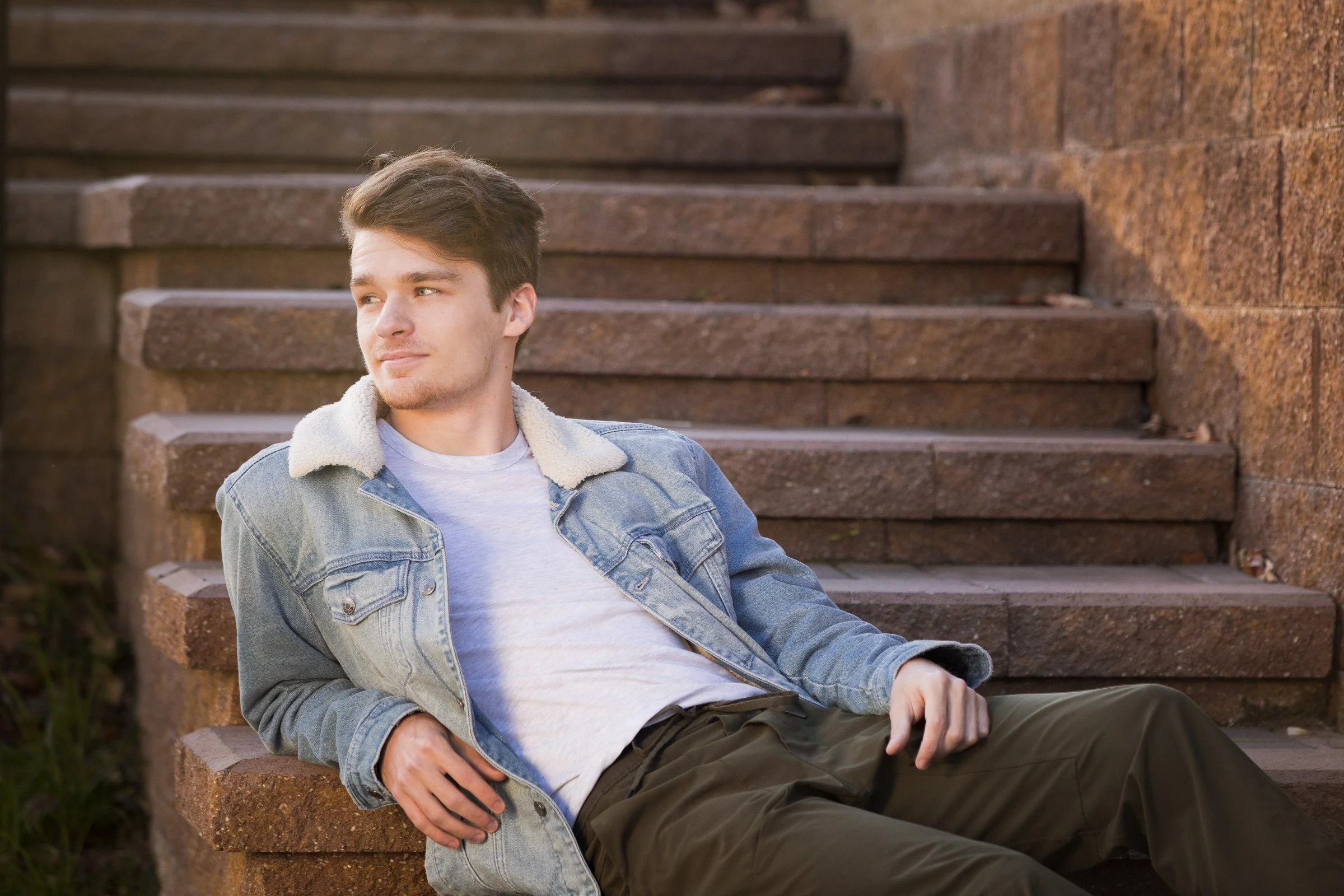 Young man wearing a denim jacket with a sherpa collar, white t-shirt, and dark pants, sitting on outdoor brick steps leaning back with his left arm resting on the step.