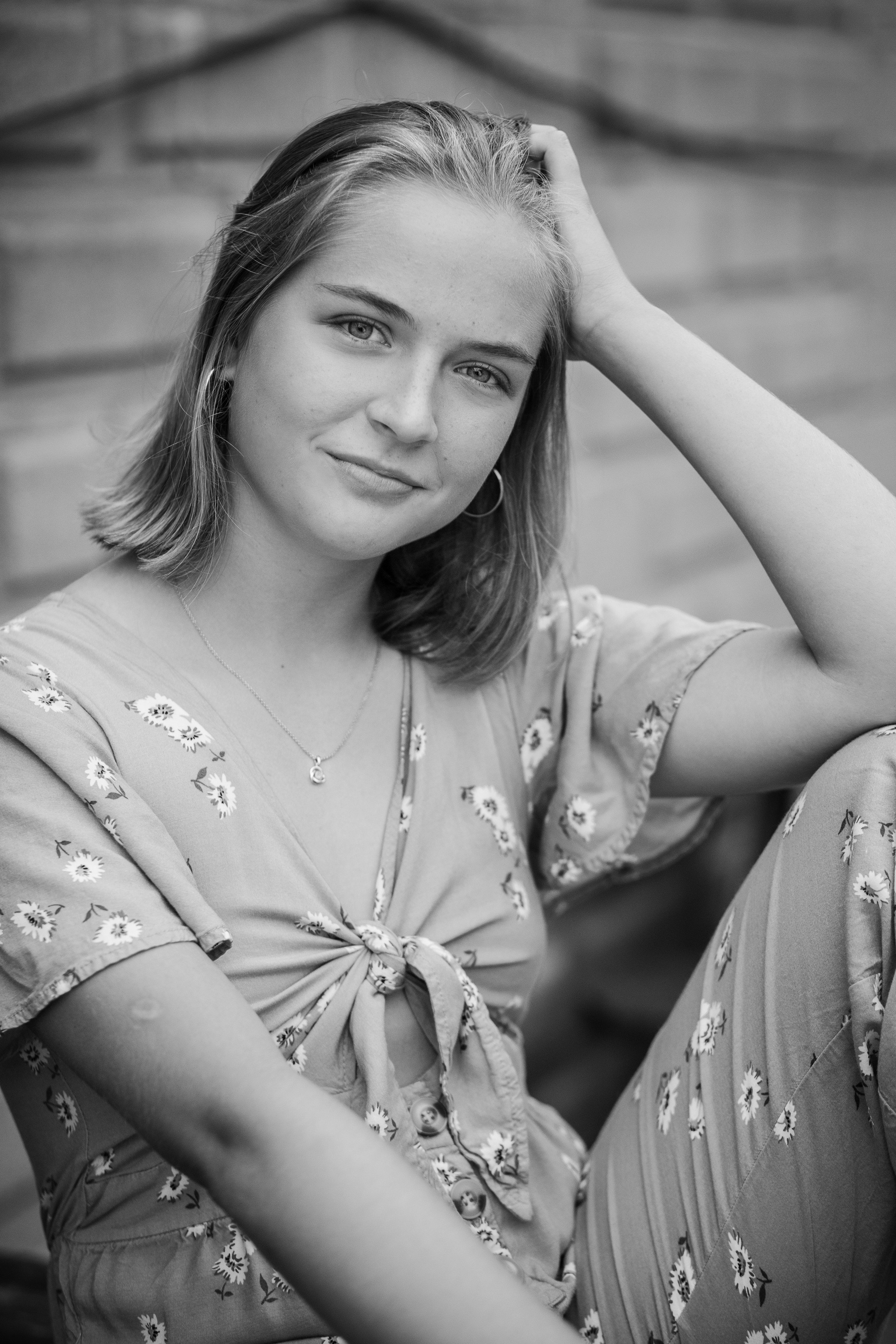Black and white portrait of a young woman with shoulder-length hair, wearing a floral dress, sitting outdoors, smiling gently at the camera.