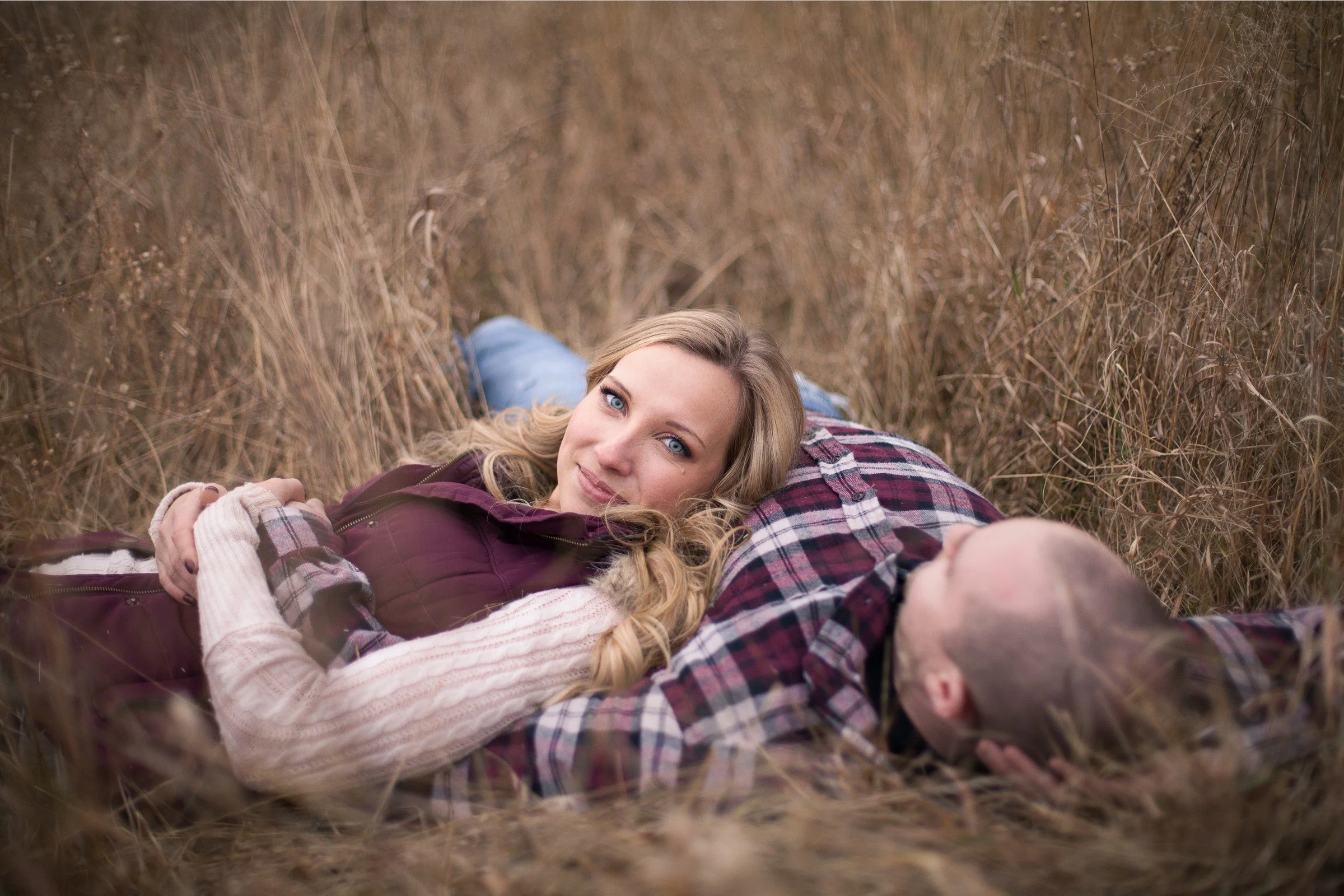 A woman with blonde hair and blue eyes lying on a man's chest in a field of tall, brown grass.