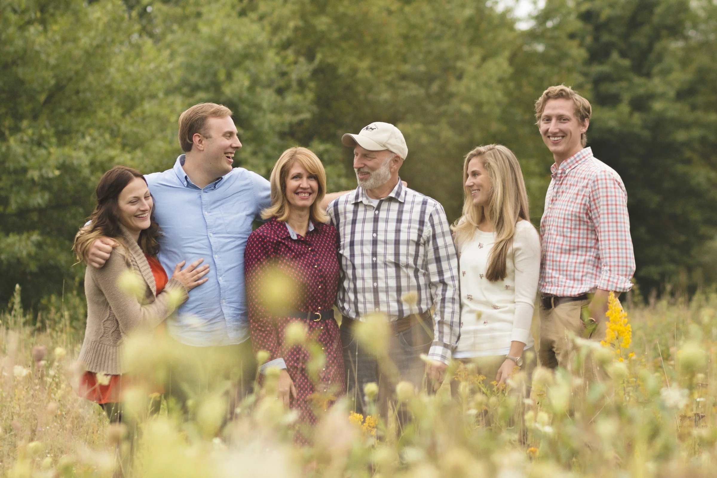 Group of six people standing in a field with greenery in the background, smiling and engaging with each other.