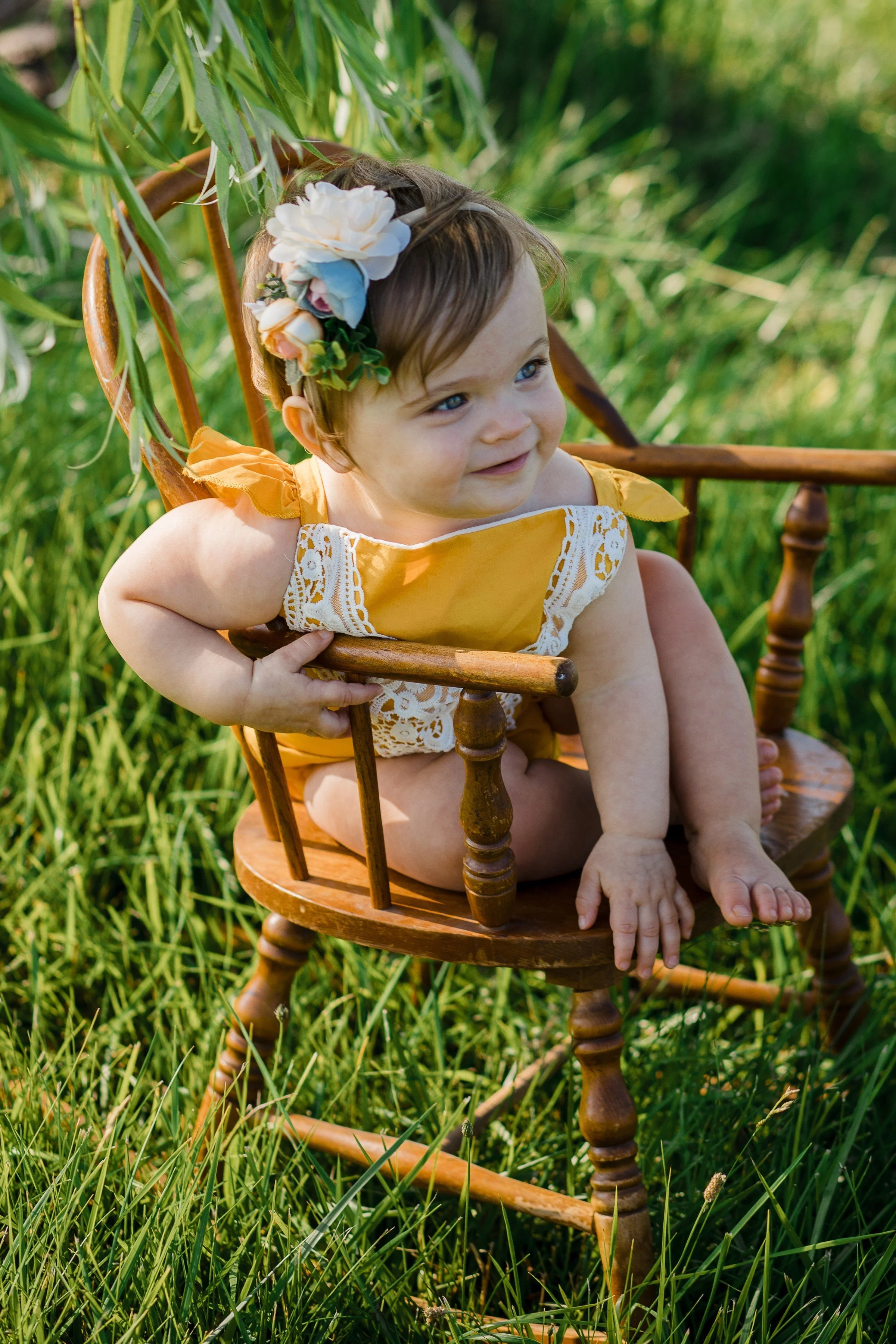 A young girl with short brown hair, wearing a yellow dress with lace trim and a flower headband, sitting on a wooden rocking chair outdoors among green grass and foliage, smiling and looking to the side.