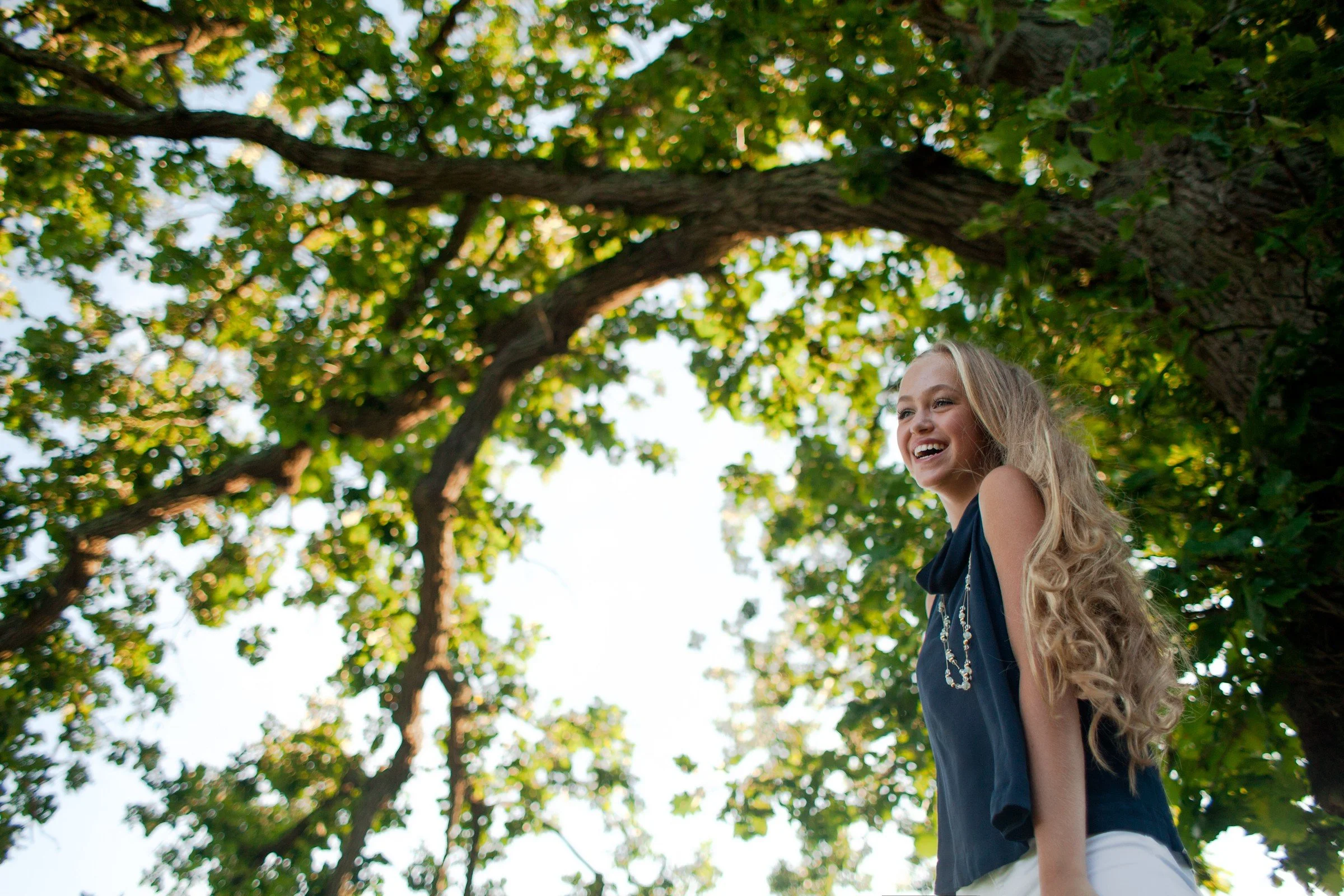 A young woman with long blonde hair smiling and standing outdoors under a large tree with green leaves.