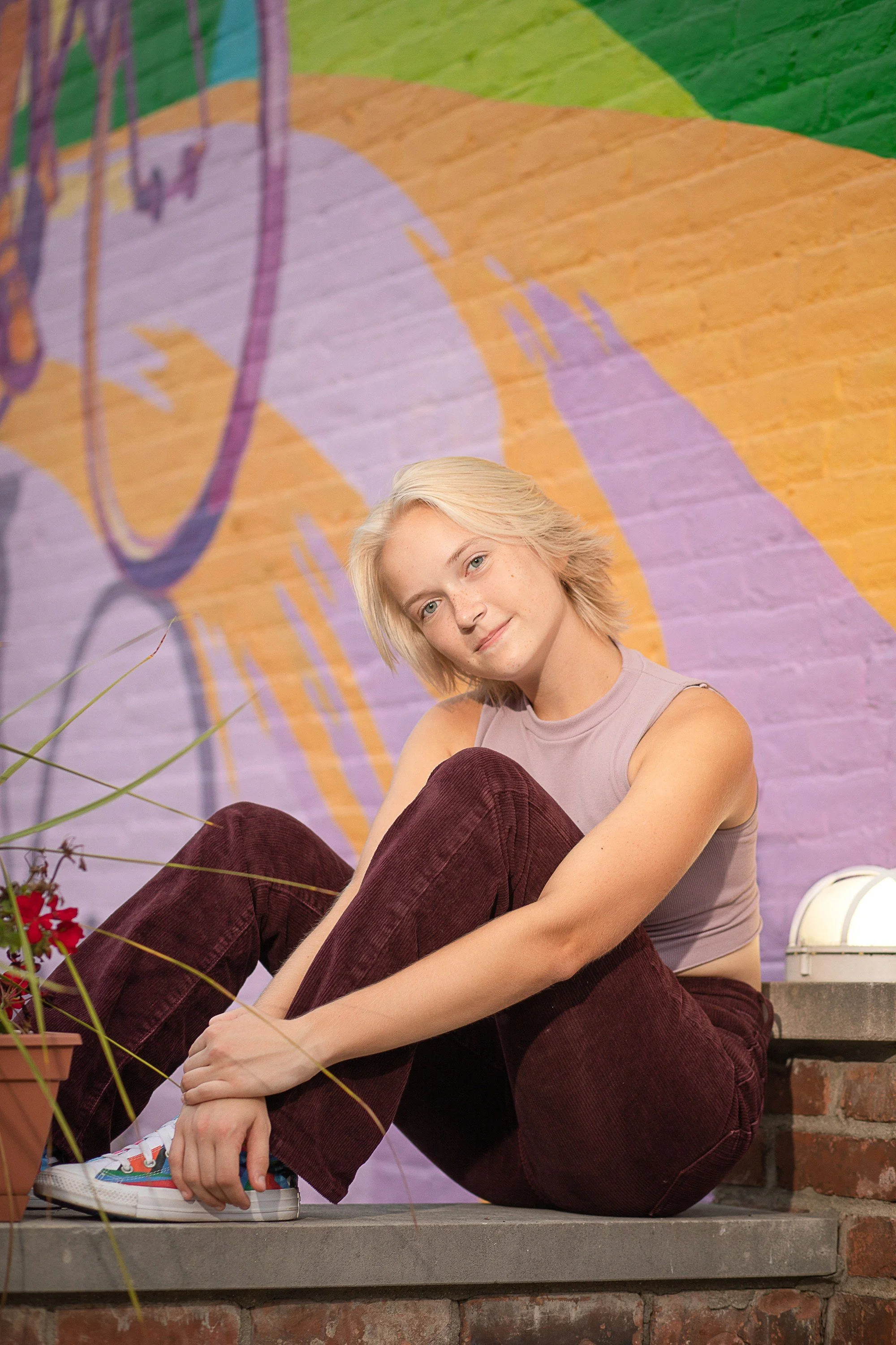 A young woman with short blonde hair sitting on a concrete step in front of a colorful mural on a brick wall, wearing a sleeveless gray top, maroon pants, and multicolored sneakers.